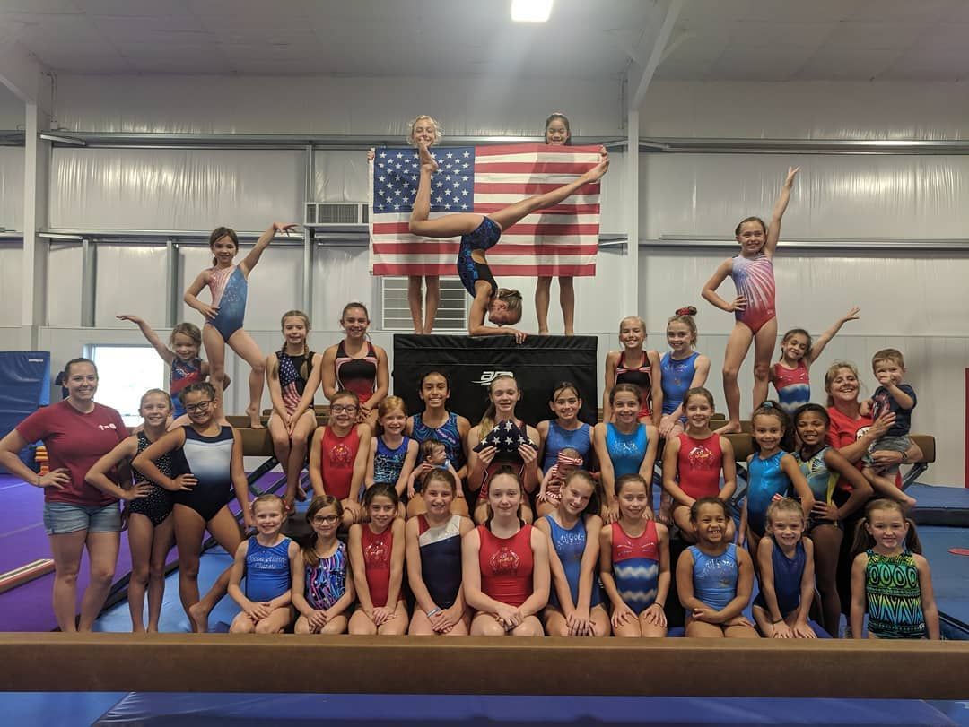 Group of gymnasts posing on a podium in a gym, with an American flag in the background.