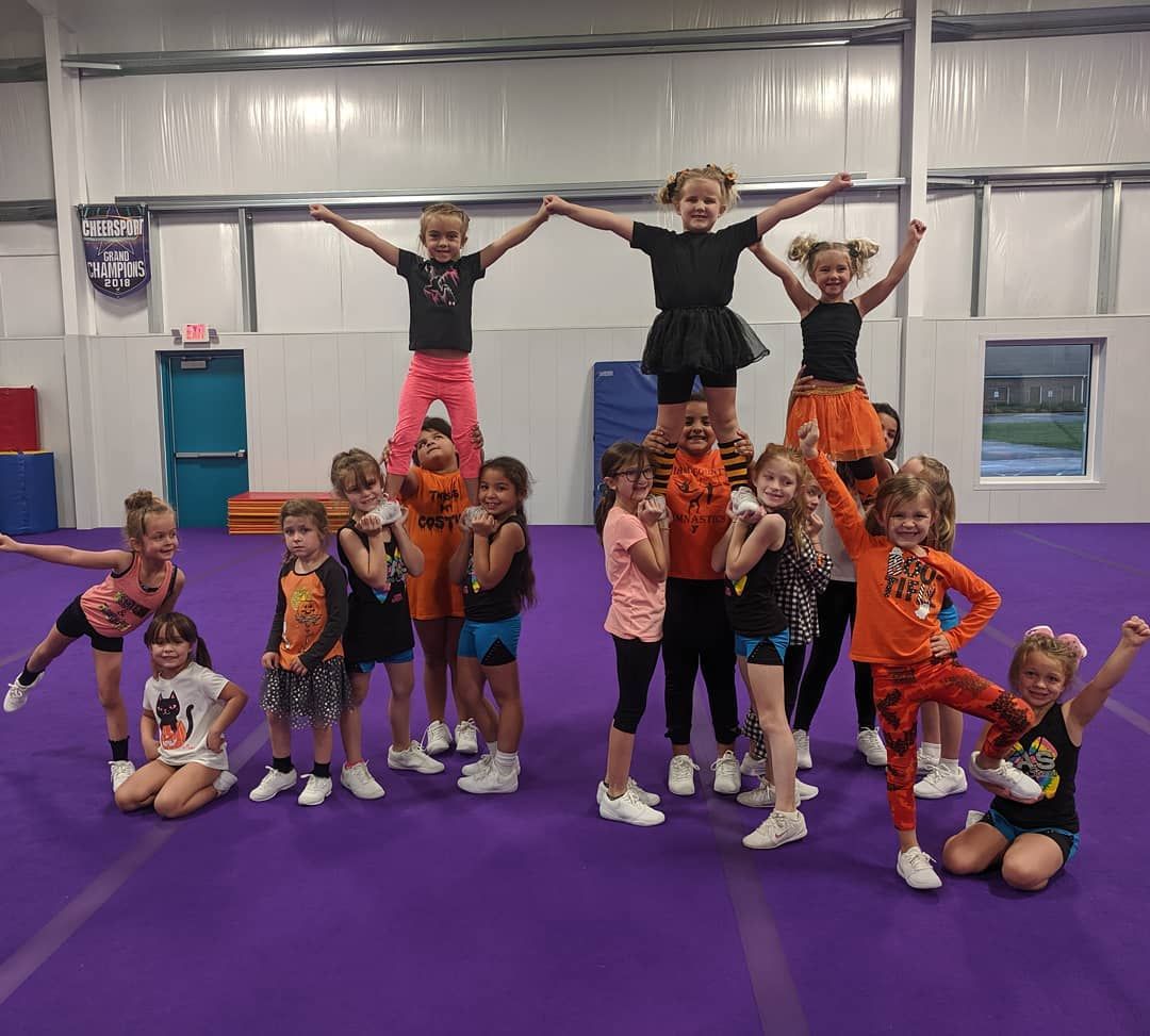 Group of children posing in cheer uniforms in a gym, some standing on others’ shoulders with arms raised.
