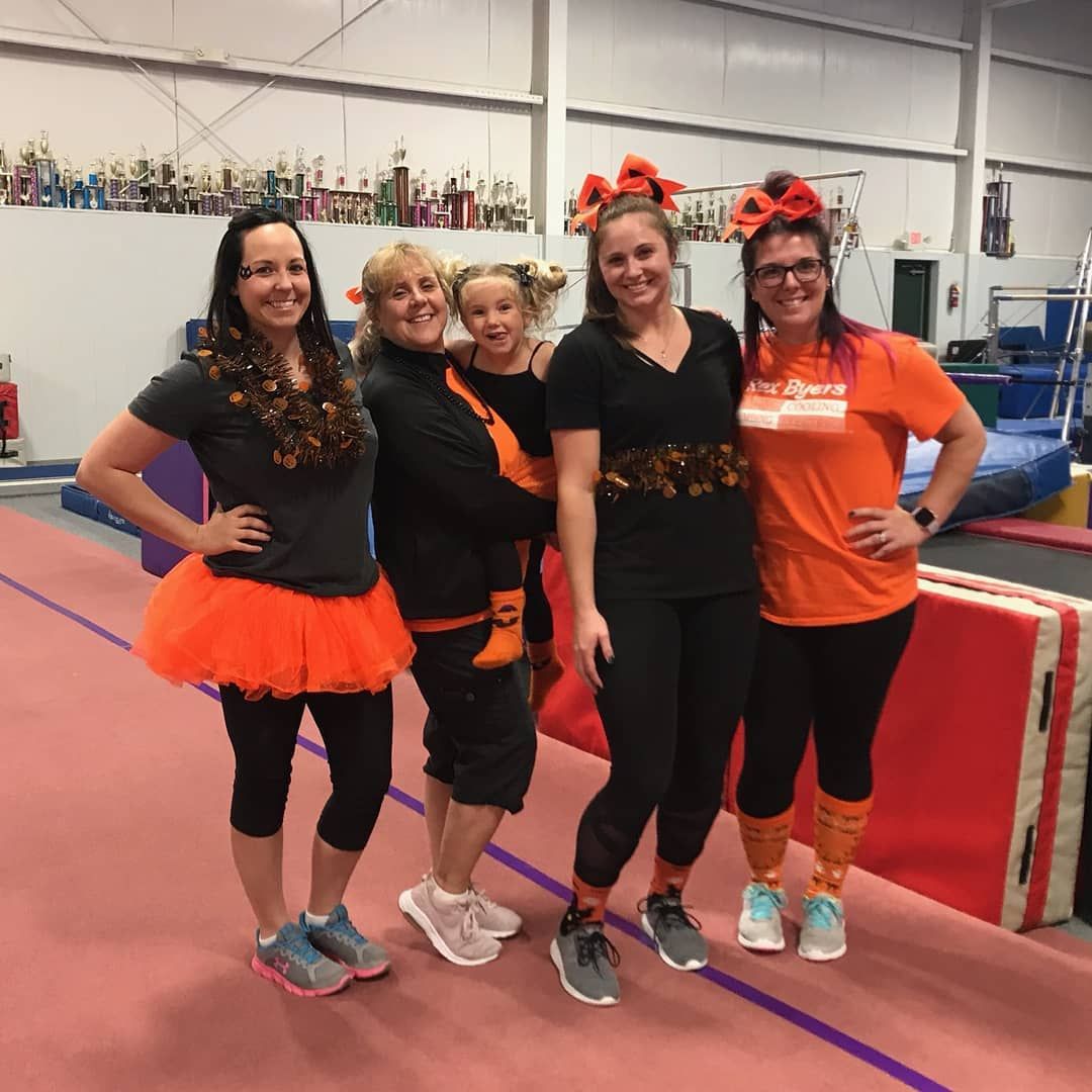Four people posing in a gymnastics gym, wearing black and orange outfits with orange bows.