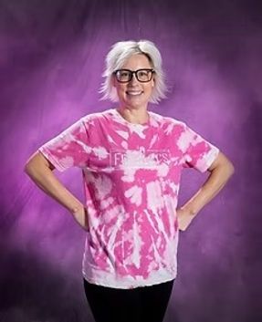 Smiling person with short white hair and glasses, hands on hips, wearing a pink tie-dye shirt against a purple backdrop