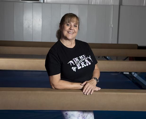 Woman in black T-shirt leaning on a wooden railing in a gym or auditorium, smiling at the camera.