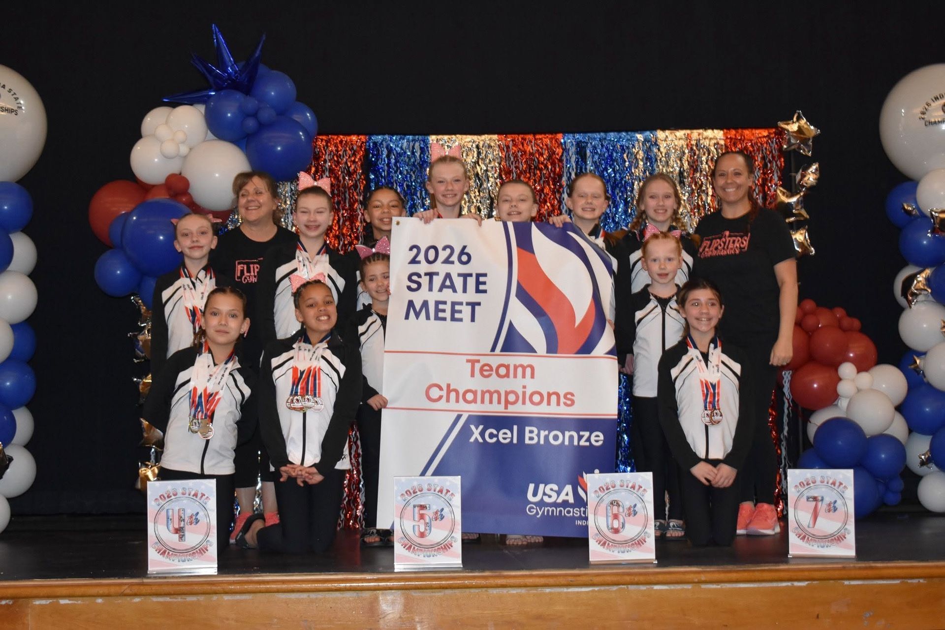 Cheer team posing on stage with “2026 State Meet Team Champions” banner and blue-white balloon decorations