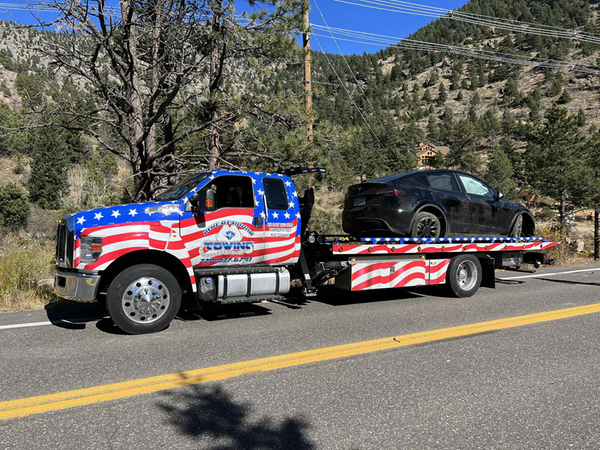 Tow truck with American flag design hauls black car on mountain road.