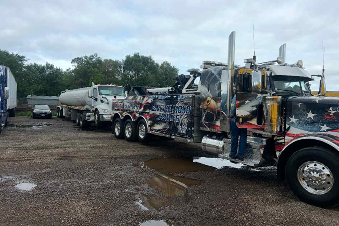 A large tow truck with an American flag design is towing a tanker truck on a gravel lot.