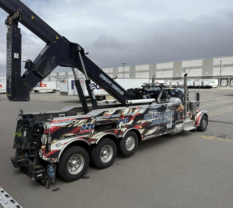 Tow truck with boom extended in parking lot, American flag design, near a warehouse.