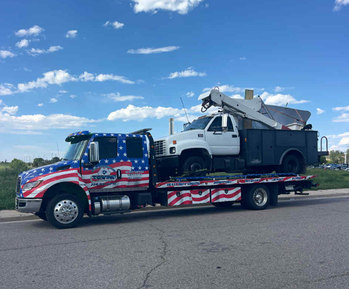 Tow truck with patriotic US flag design, carrying a white truck under a blue sky.