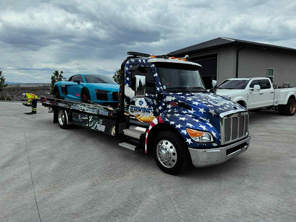 Tow truck with blue patriotic stars and stripes hauling a blue sports car, parked on a paved driveway.