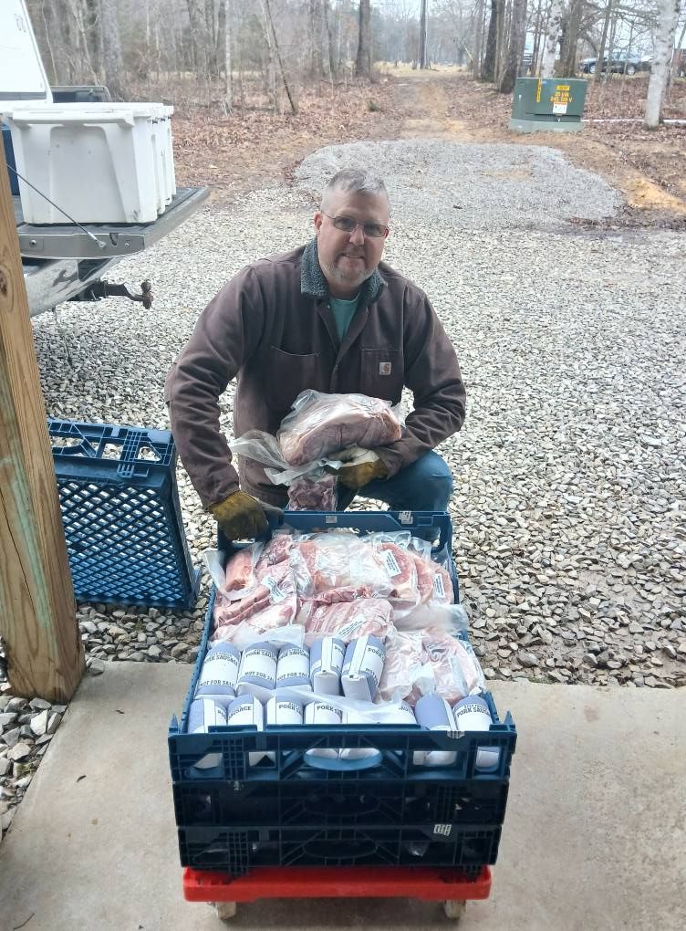 Man with meat packages in a crate, outside. He smiles, wearing work clothes. Concrete and trees in background.