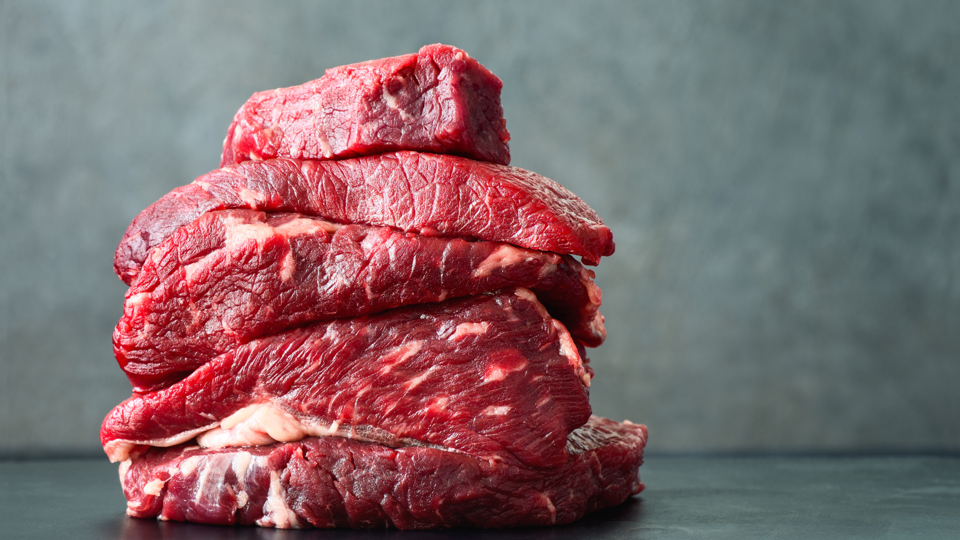 Stack of raw, red meat steaks on a dark surface.