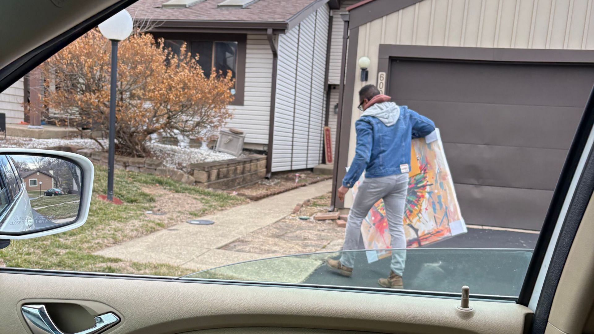 Person carrying a large framed item near a driveway outside a house, seen from a car.