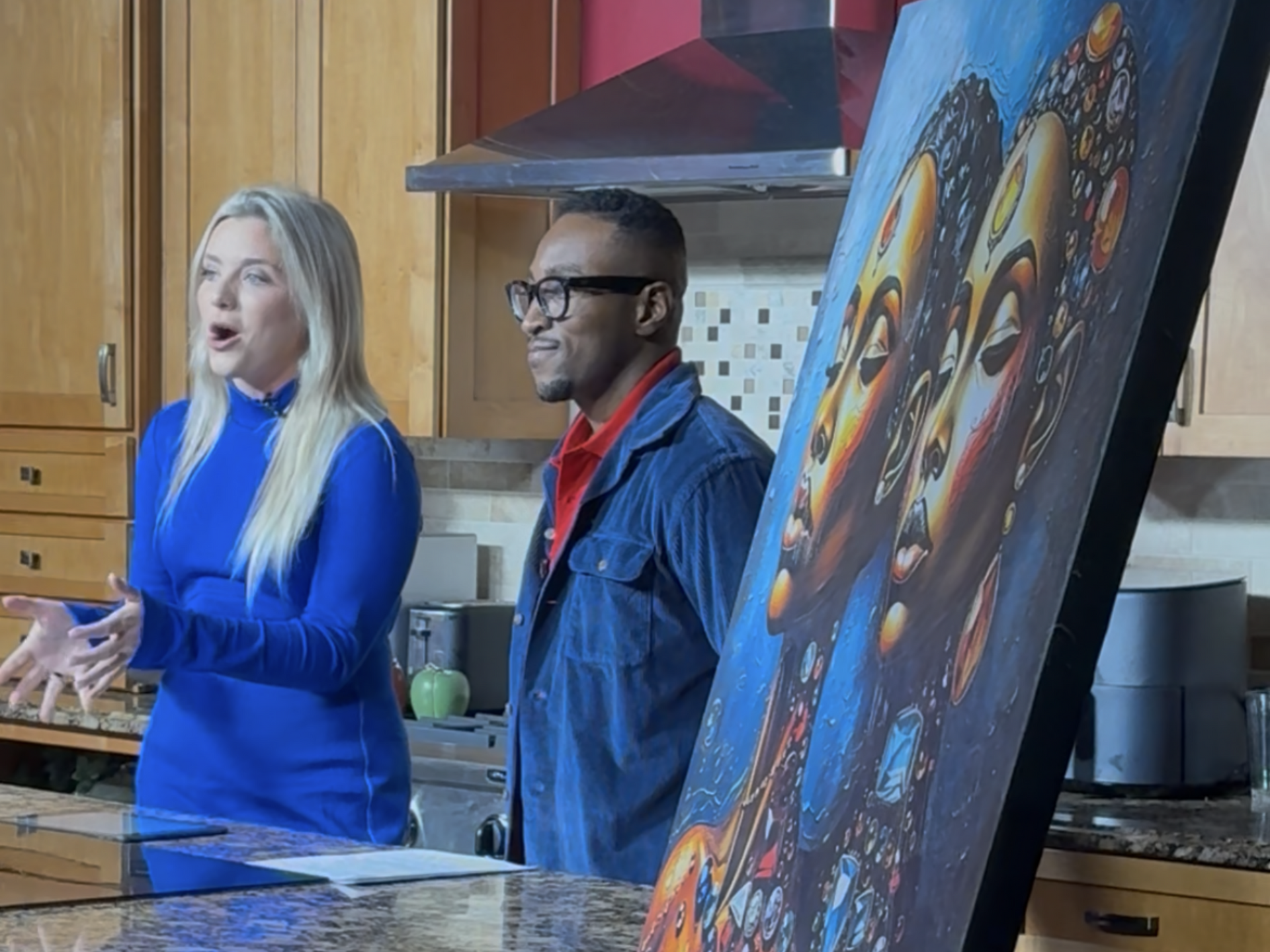 Two people speaking in a kitchen beside a large colorful portrait painting