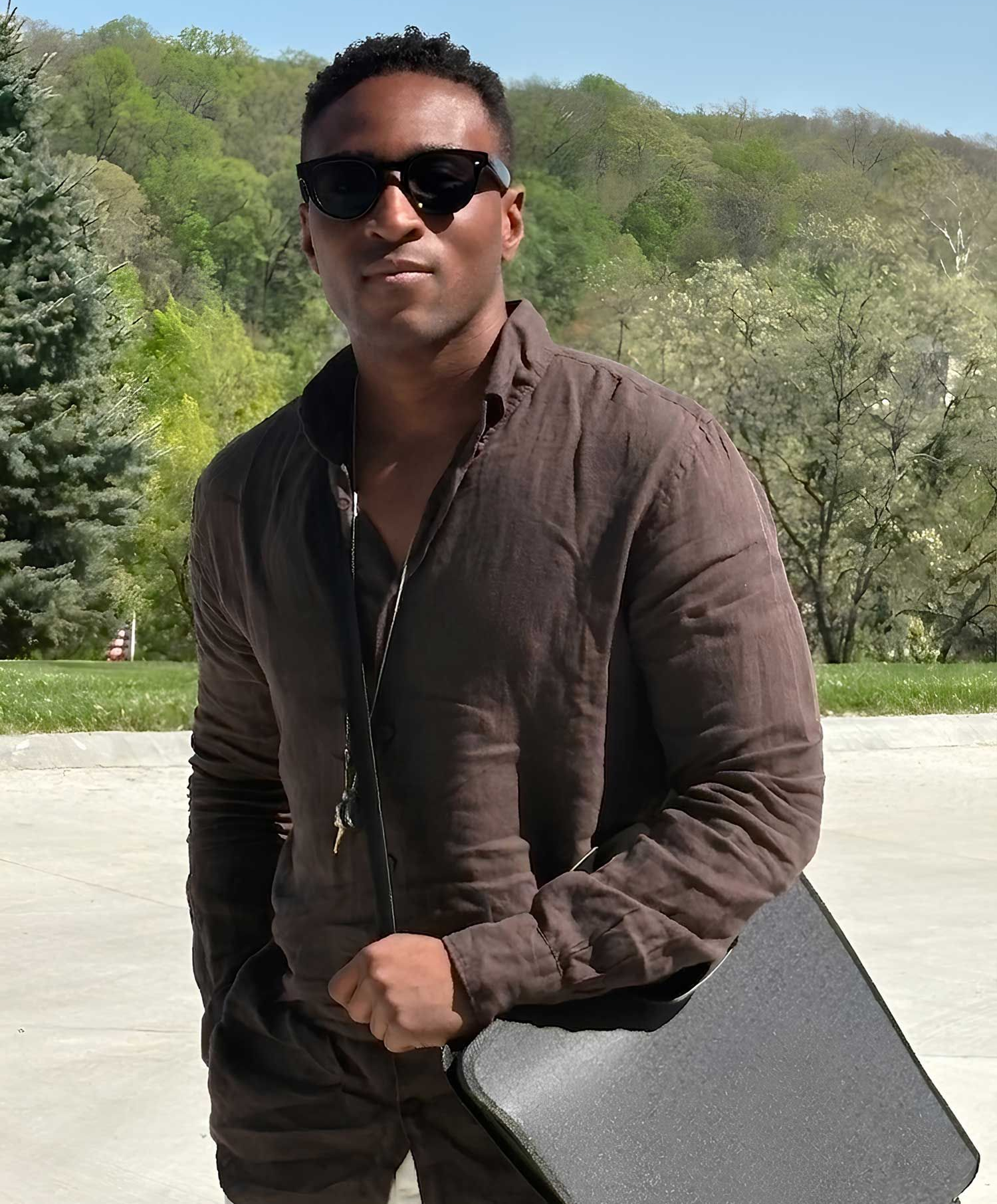Man in sunglasses holding a black briefcase outdoors in a park-like setting