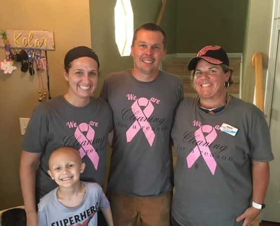 Family of four standing indoors, smiling, wearing matching gray shirts with pink breast cancer ribbons.