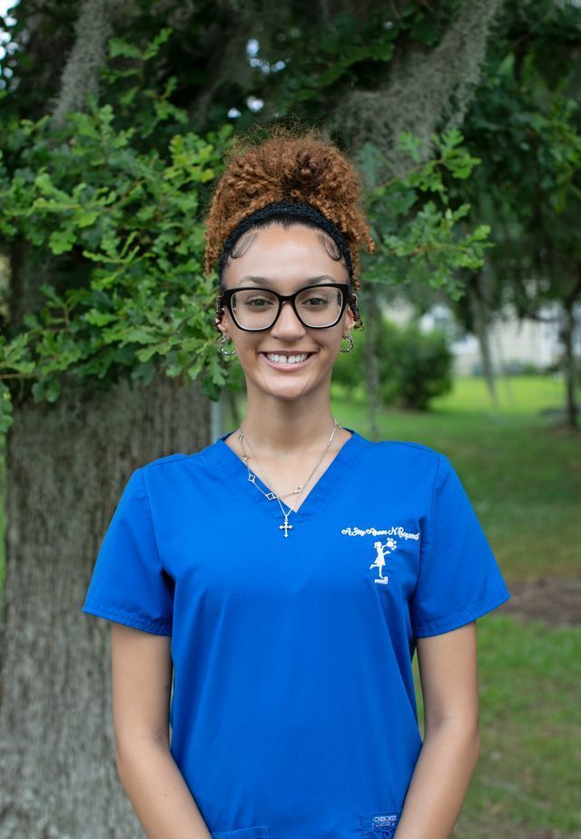 Smiling person in a blue shirt standing outdoors by a tree and greenery