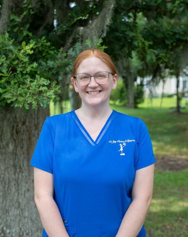 Smiling person in a blue shirt standing outdoors by green trees