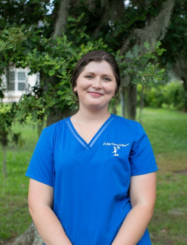 Woman in blue scrubs smiling, in a car. Scrubs have a logo. Brown hair, light skin.