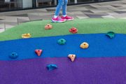 Child standing on colorful climbing playground with multicolored holds and textured purple, blue, and green surfaces