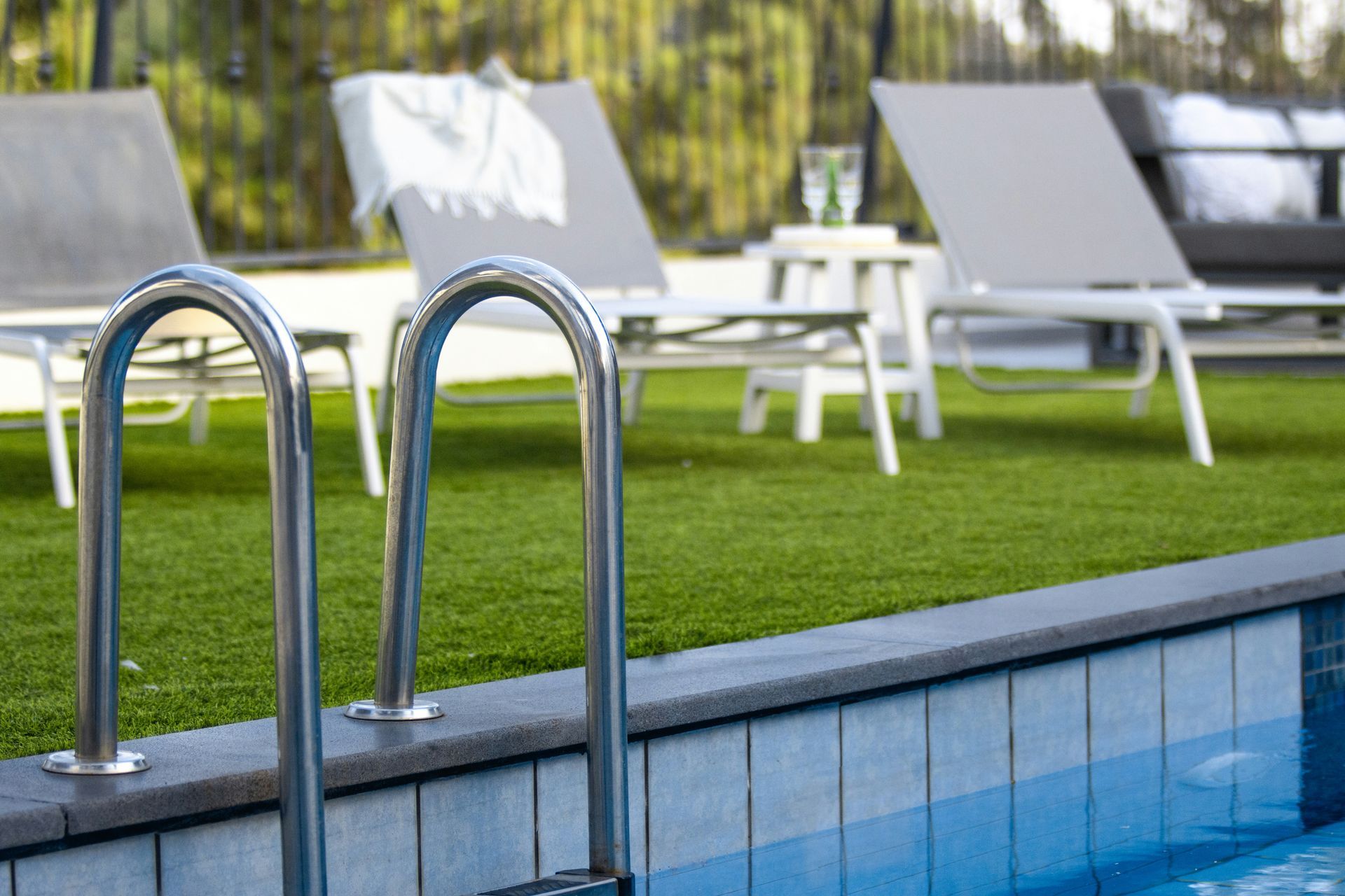Pool ladder beside a blue-tiled pool with white lounge chairs on a green lawn.