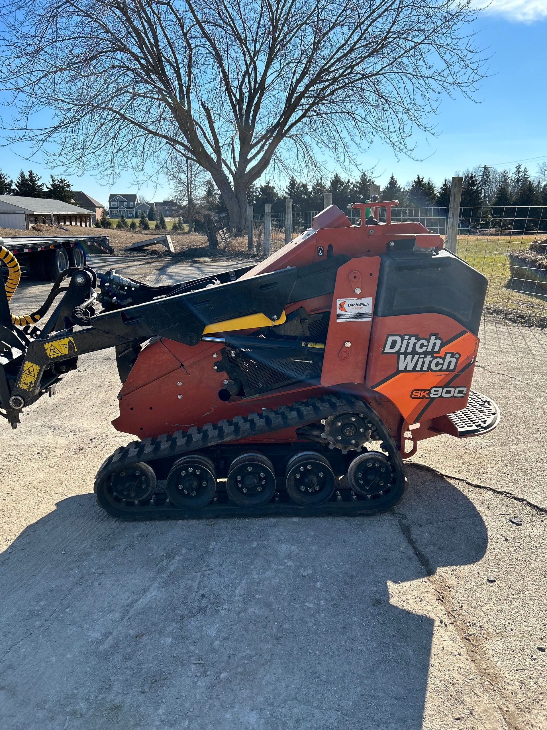 Orange Ditch Witch mini skid steer with tracks and arm in a gravel yard under a leafless tree