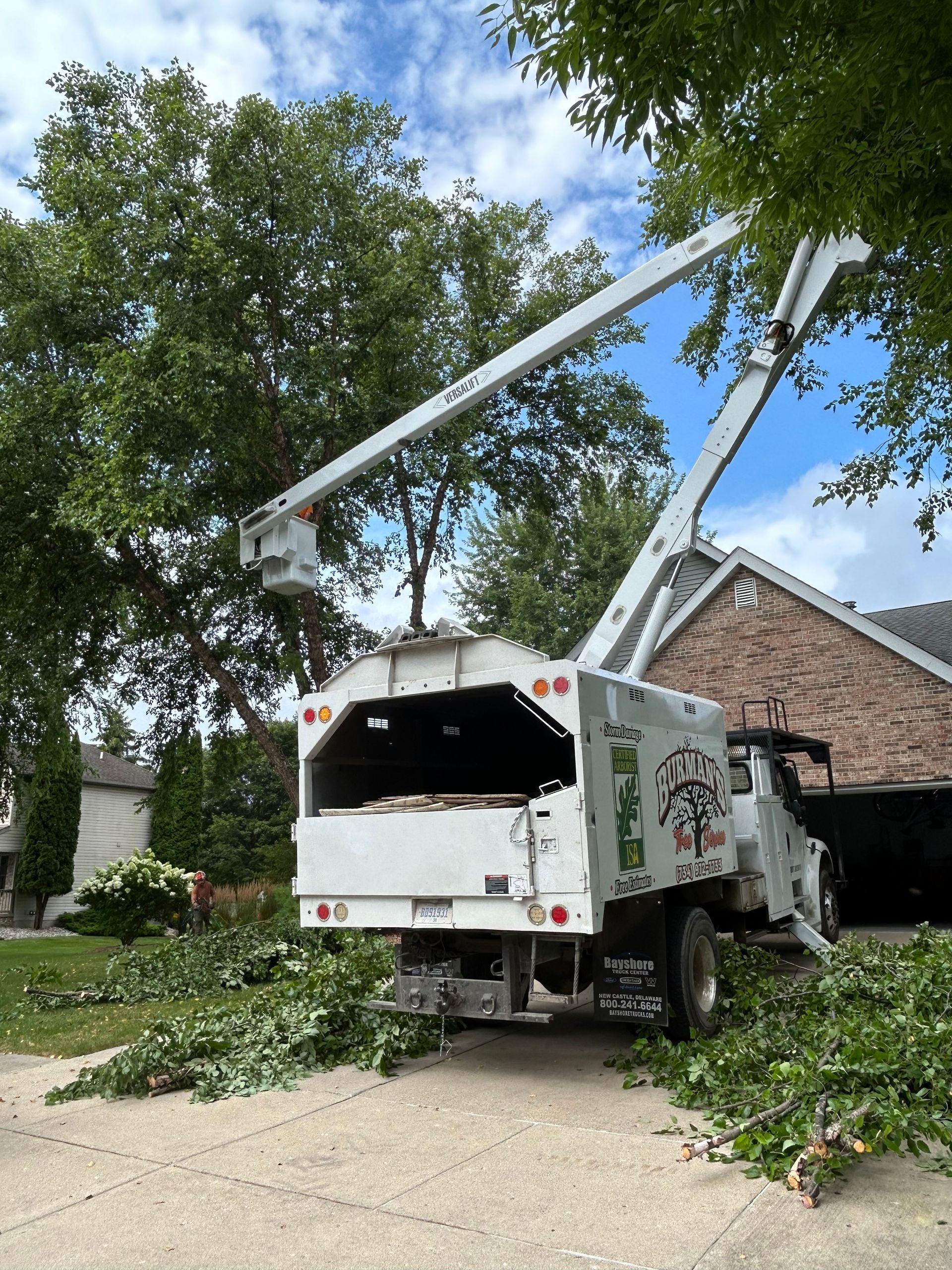 Utility truck trimming tree branches on a residential street, with limbs and leaves piled on the driveway.