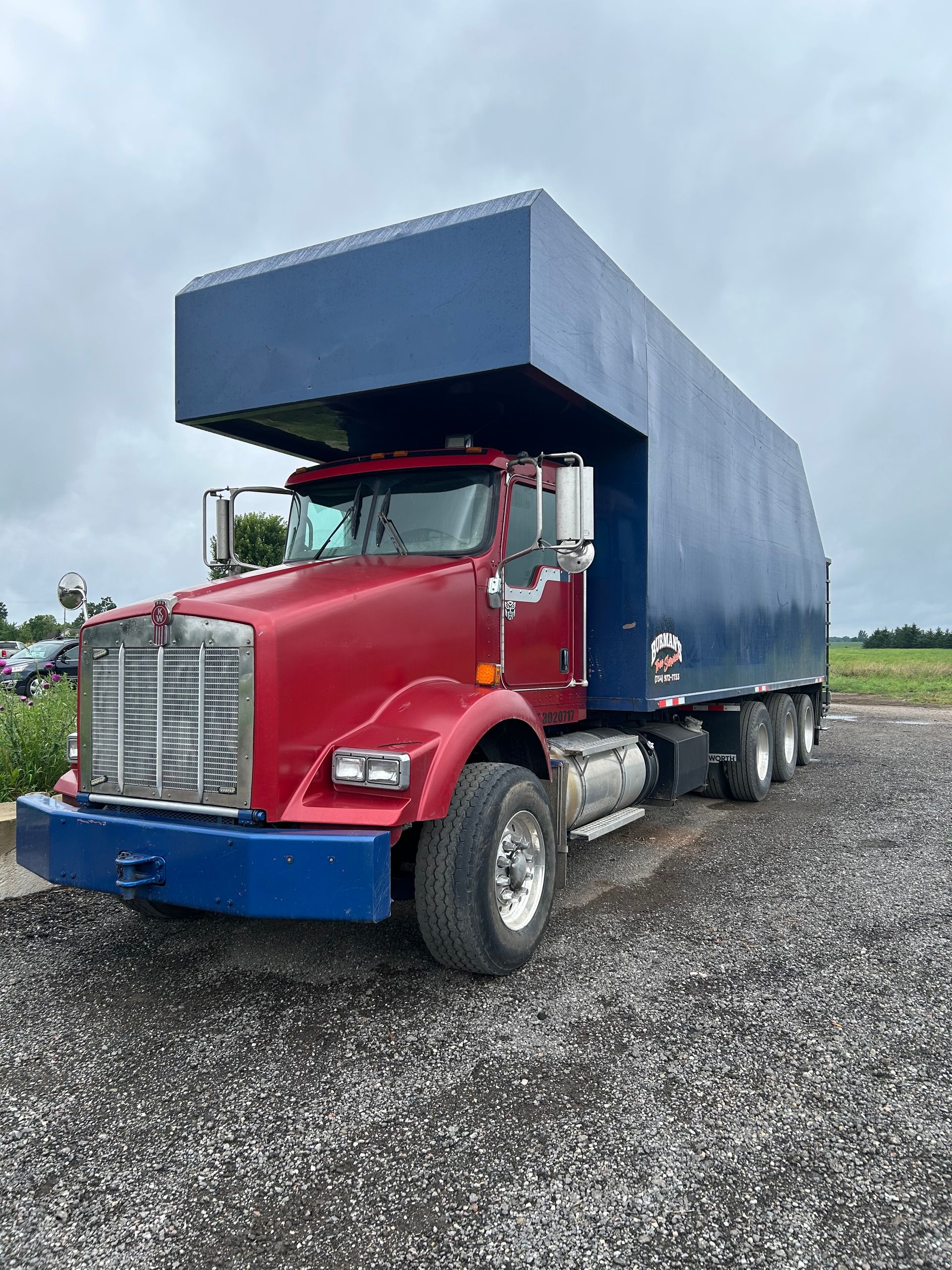 Red semi-truck with blue cargo box parked on gravel under a cloudy sky