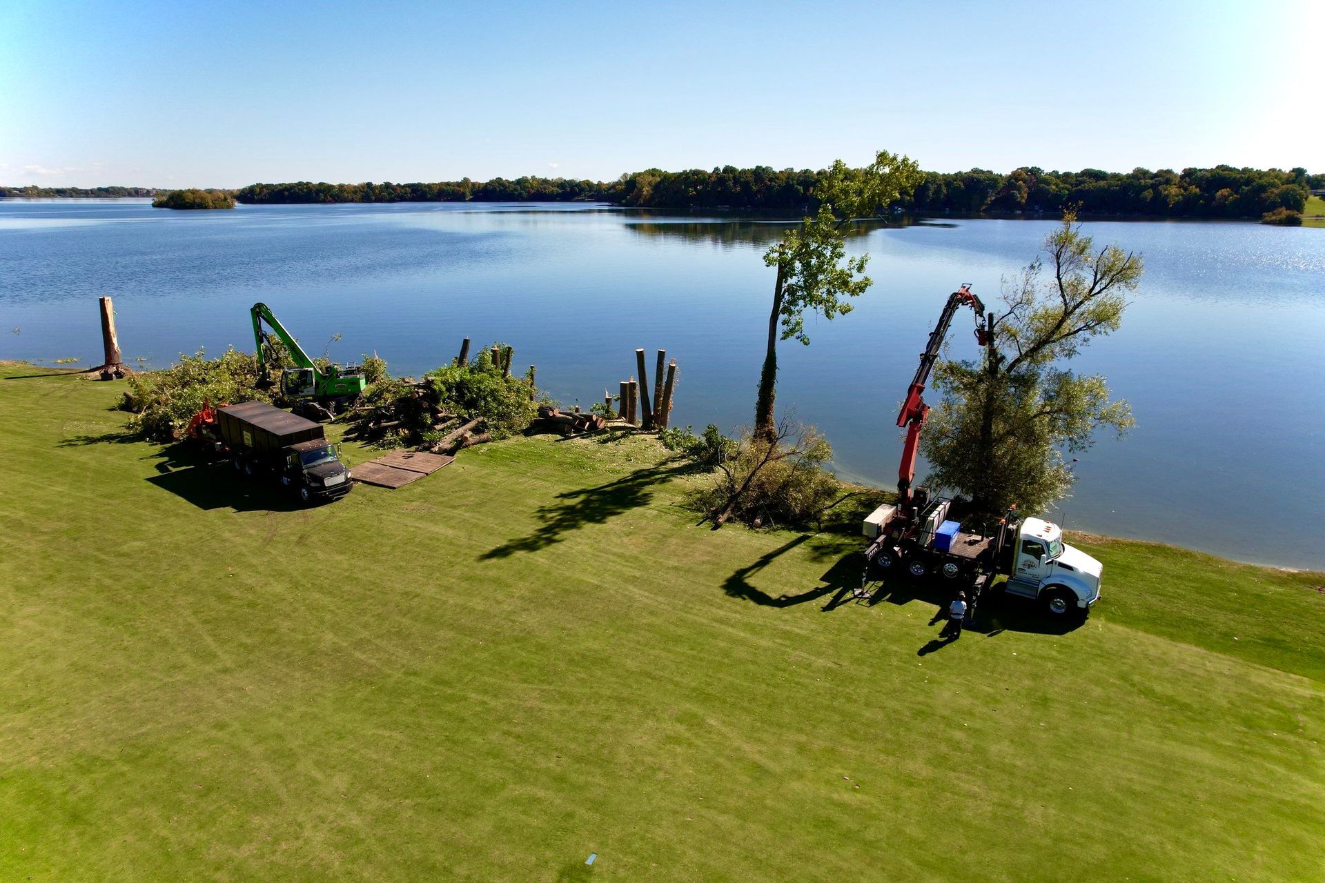 Lawn beside a calm lake with a flowering tree, patio chairs, and a distant shoreline under a clear blue sky