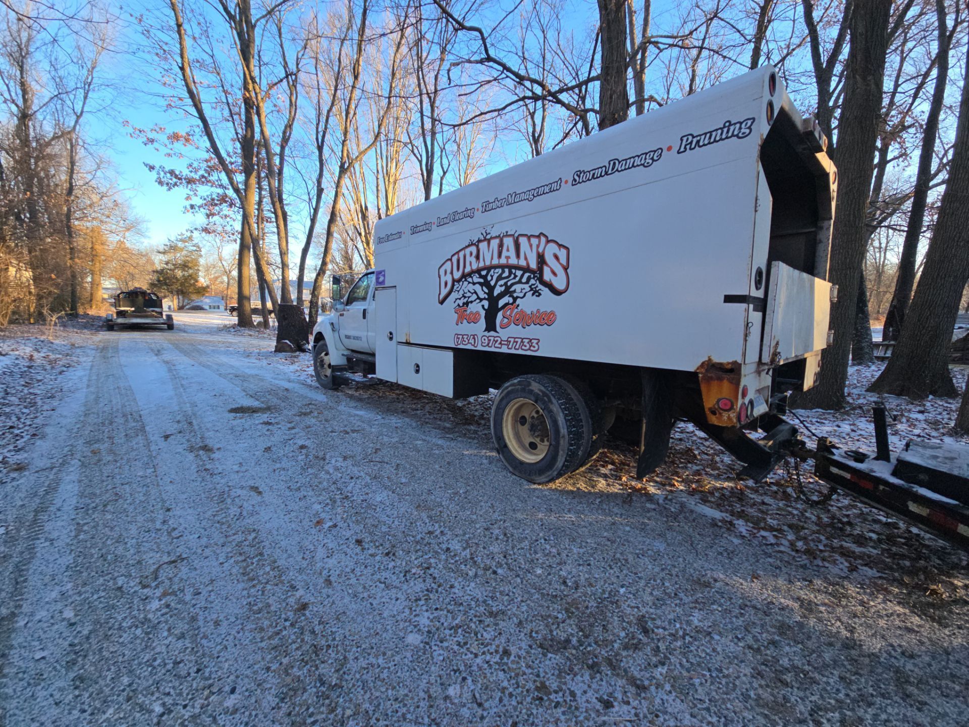 White delivery truck tipped off a snowy road beside trees, with a damaged rear wheel.