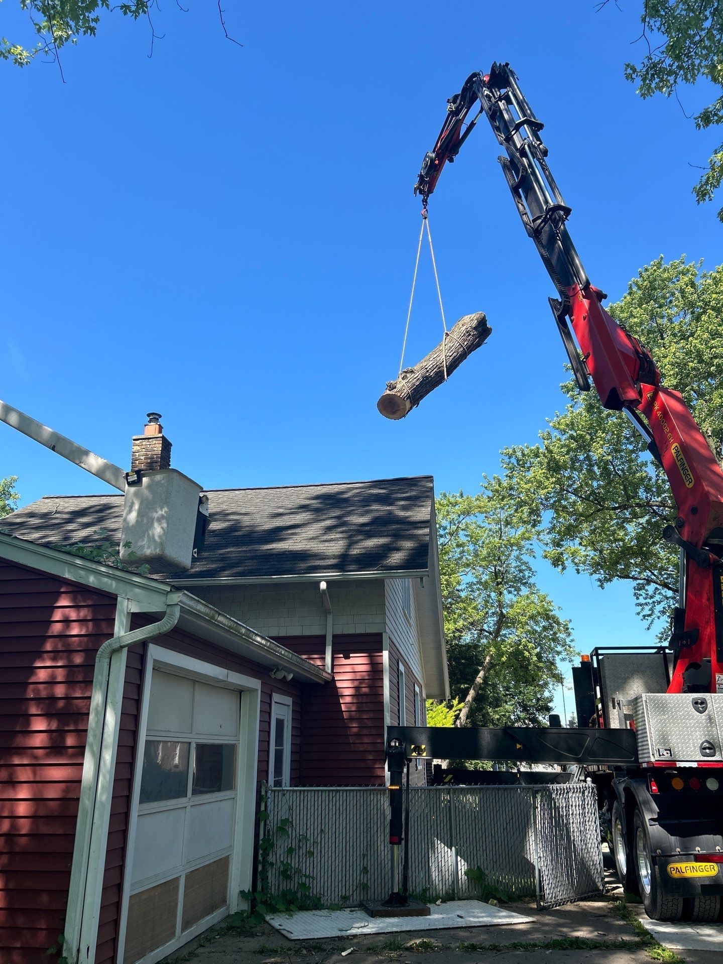 Crane lifting a large log over a red house and fenced yard under a clear blue sky