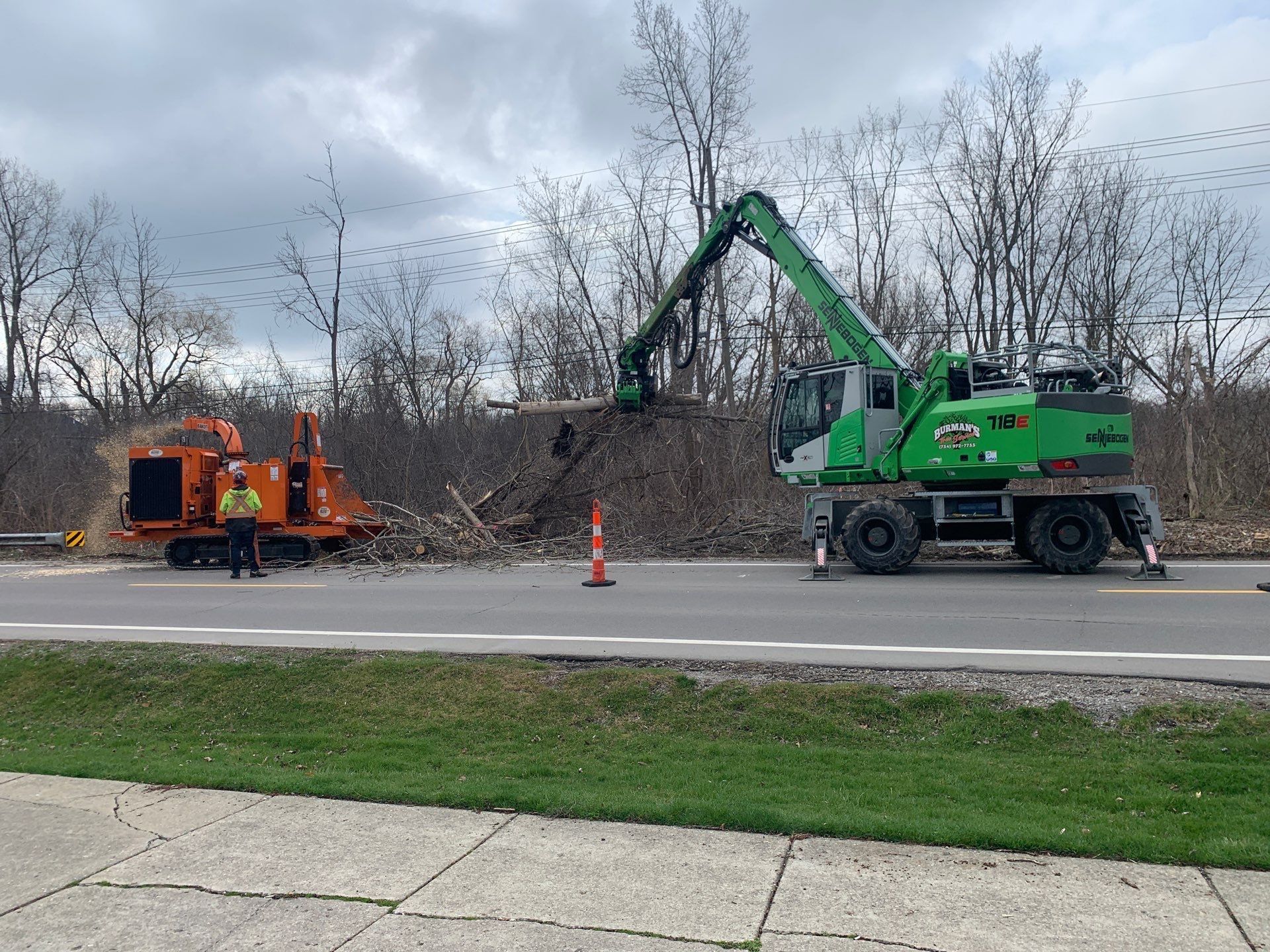 Green utility truck and orange road machine working beside a roadside, with cones and bare trees.