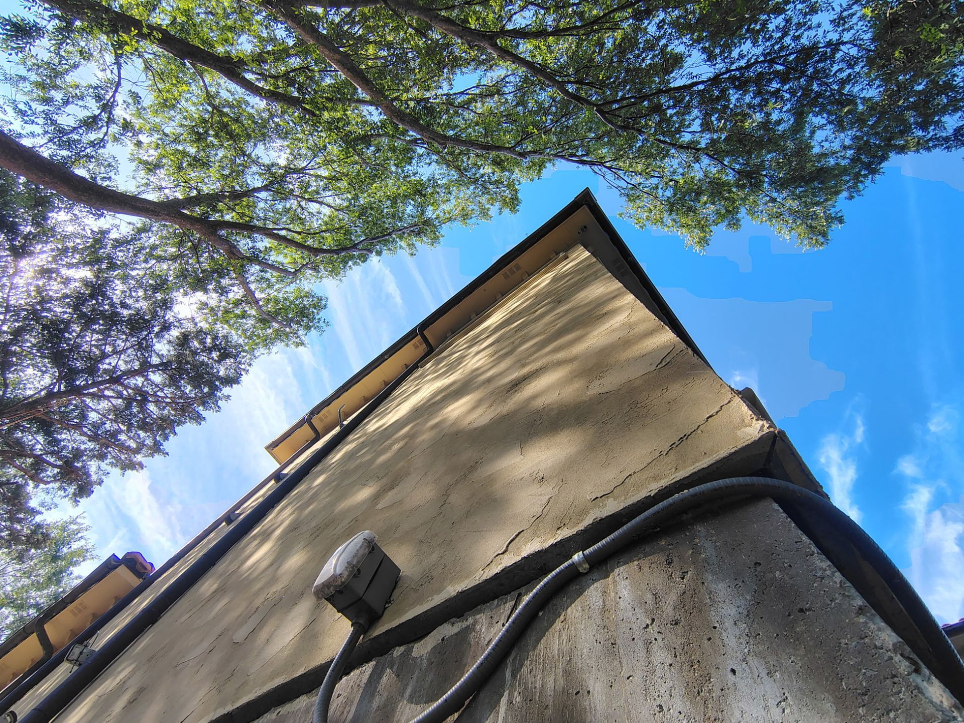 Low-angle view of a beige building corner against a blue sky, framed by tree branches and cables