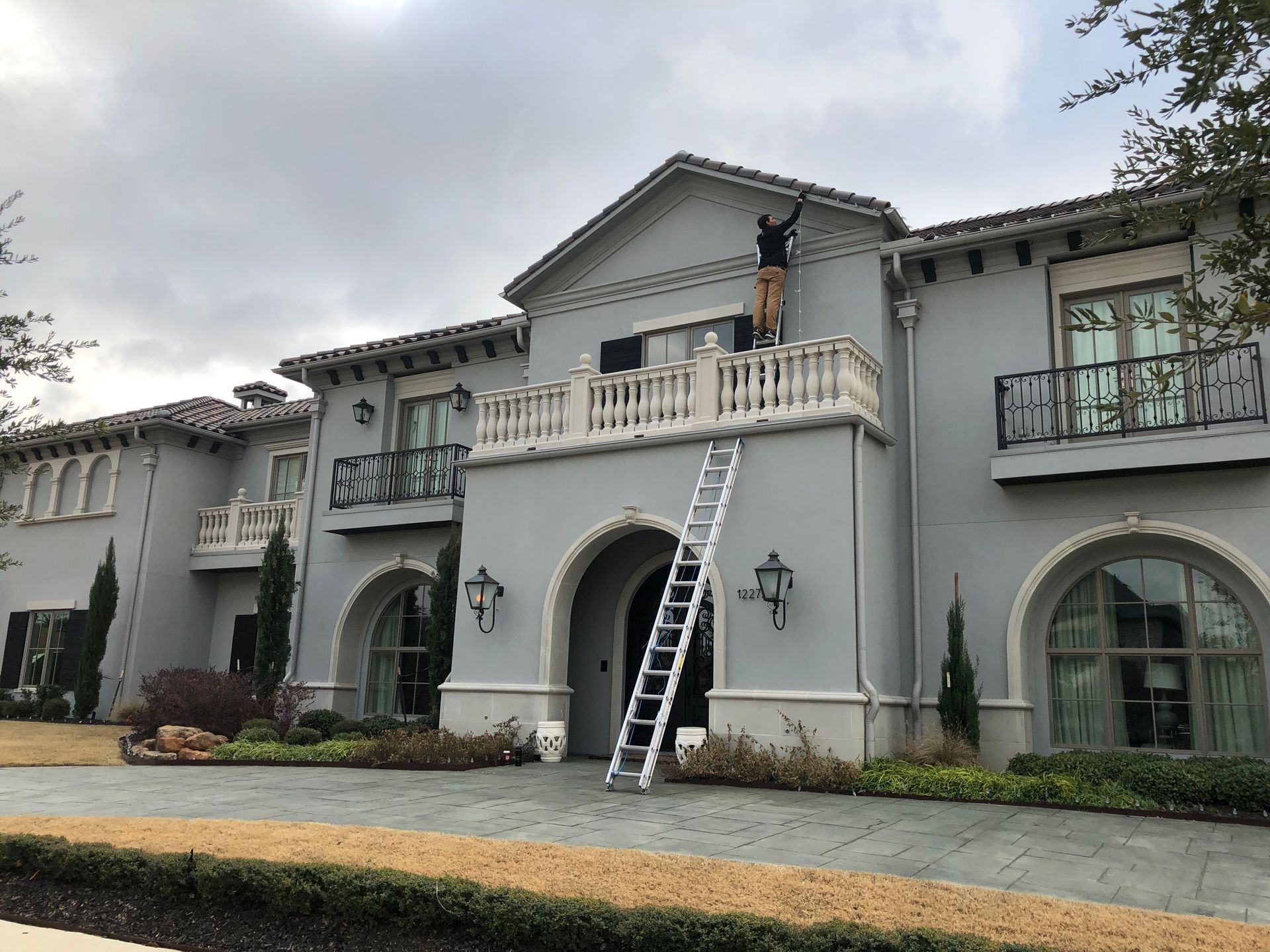 Large white stucco house with balconies; a ladder leans to the upper balcony where a person is working.