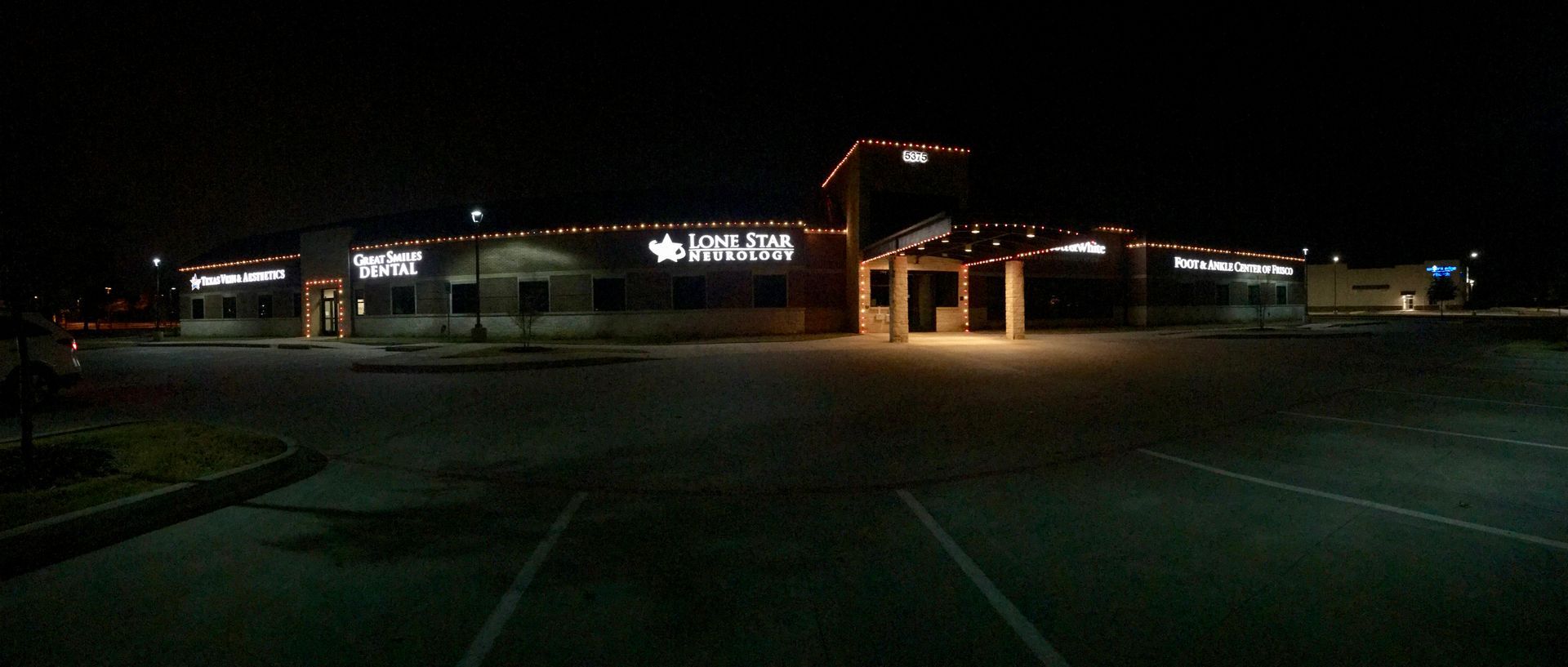 A dark nighttime view of a lit building and parking lot, with lights glowing under an entrance canopy.