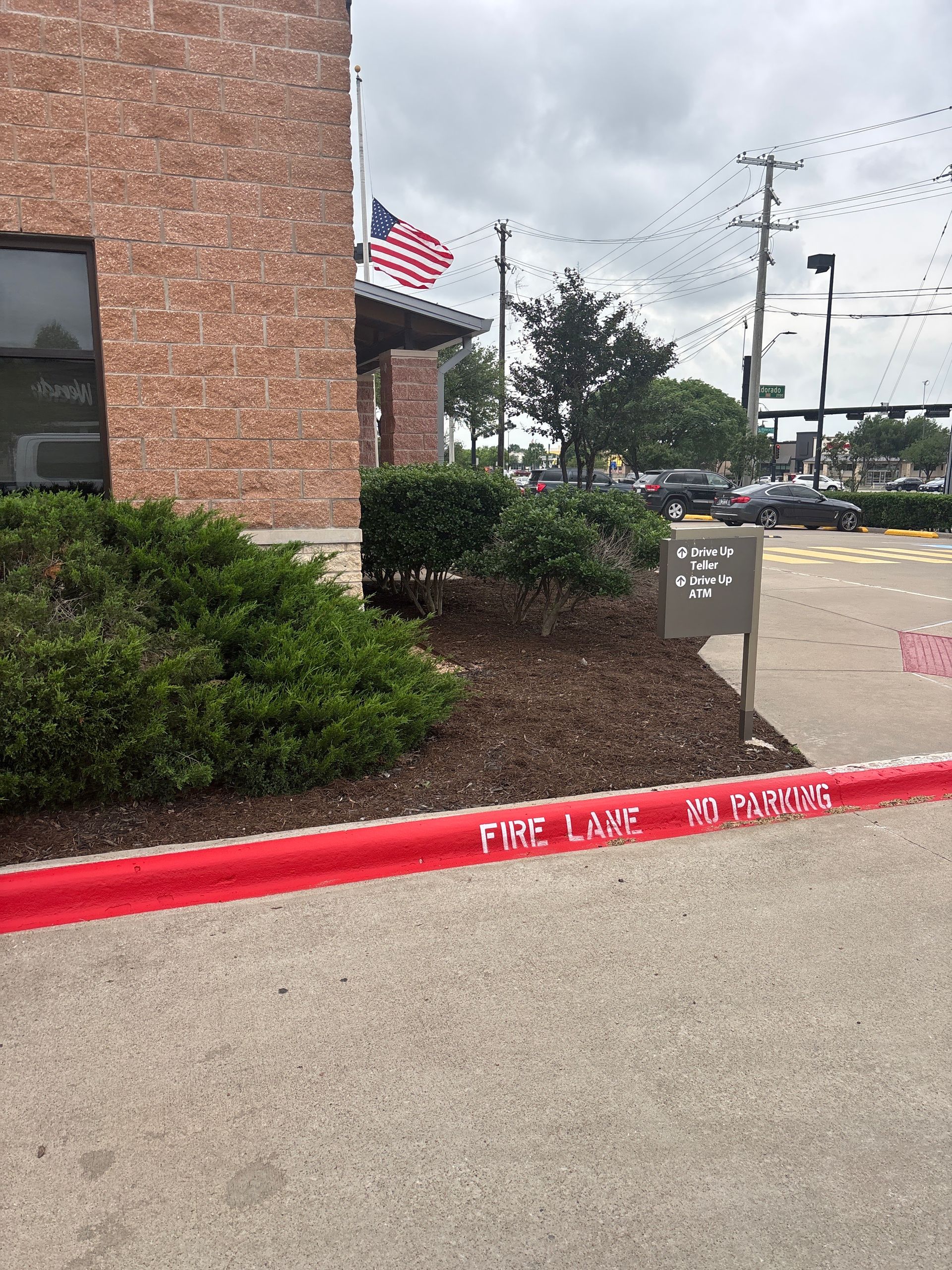 Brick building corner with shrubs, red curb, and American flag near a parking lot entrance.
