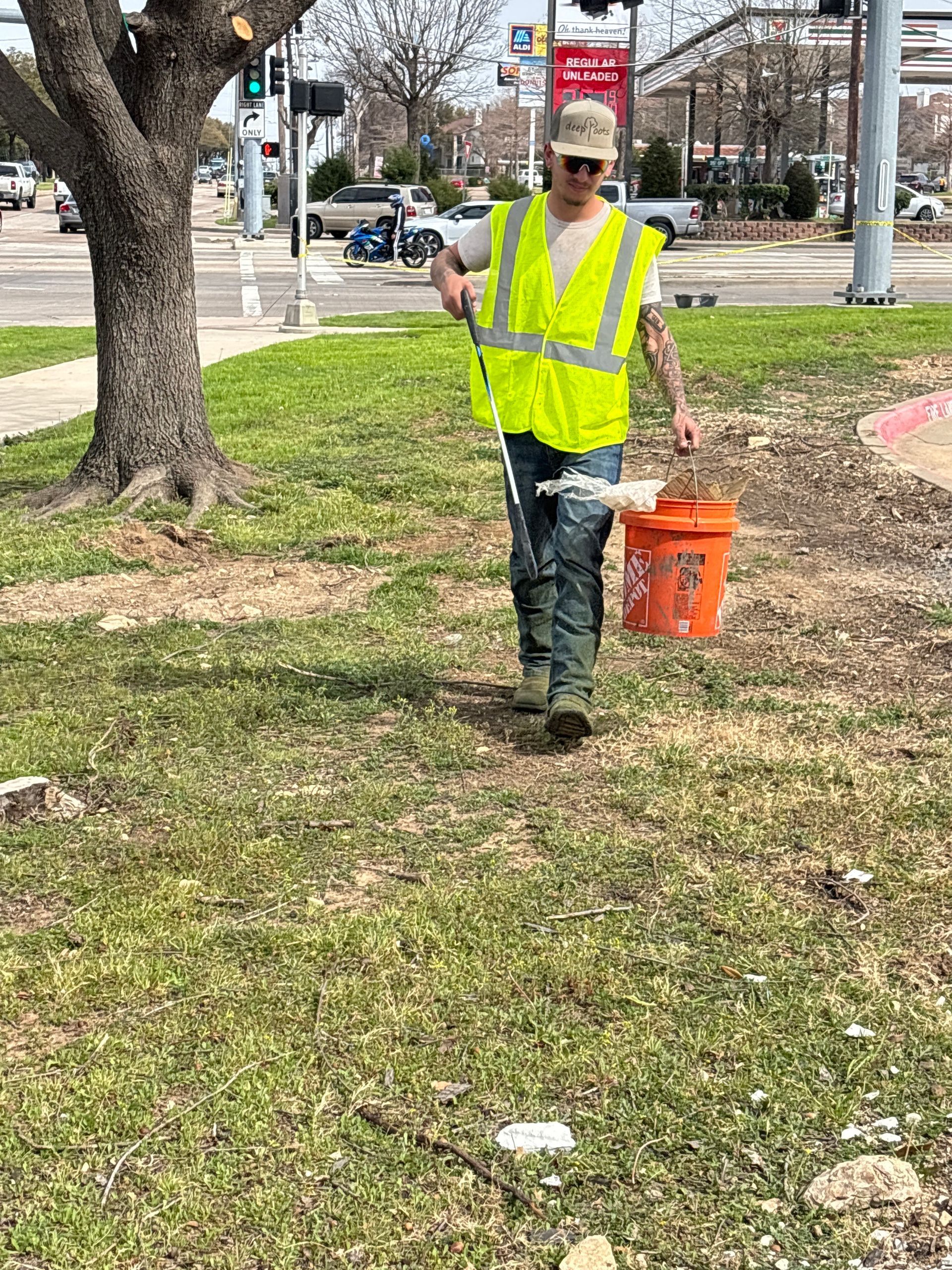 Worker in neon safety vest walking on grass with an orange bucket in a park.