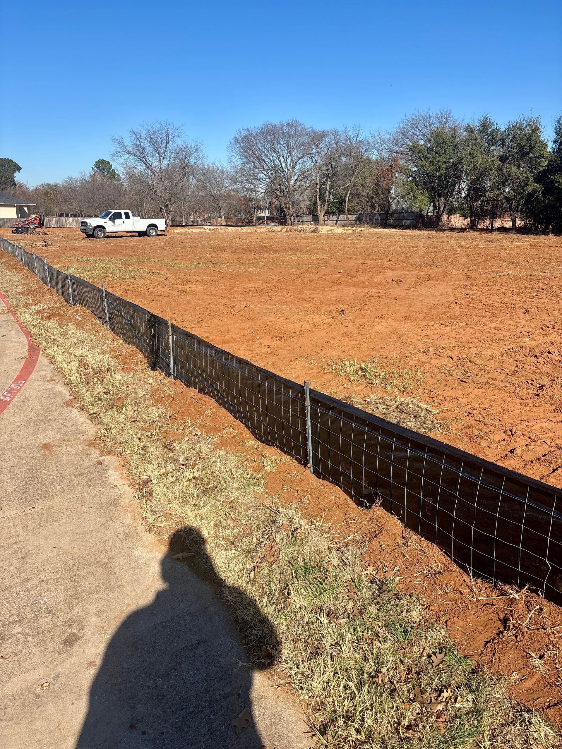 Dirt lot with black silt fence beside a path, white truck in the distance, and bare trees under a clear blue sky