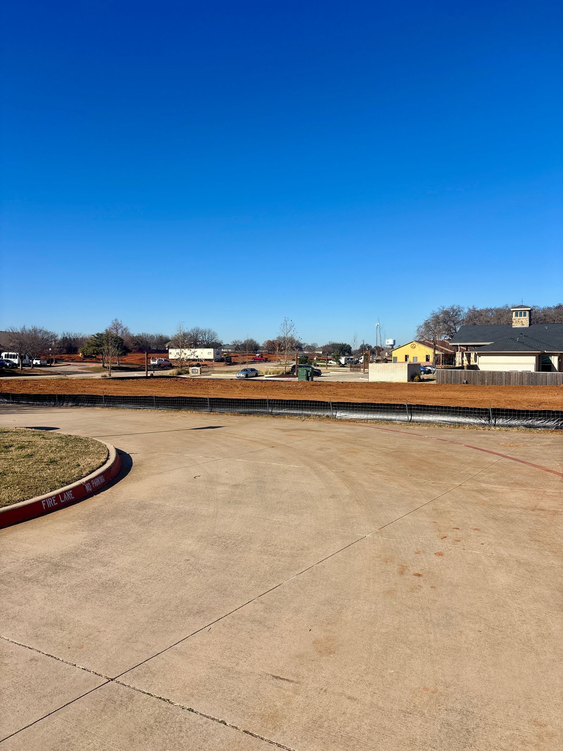 Empty paved lot under a clear blue sky, with low buildings and trees in the distance.