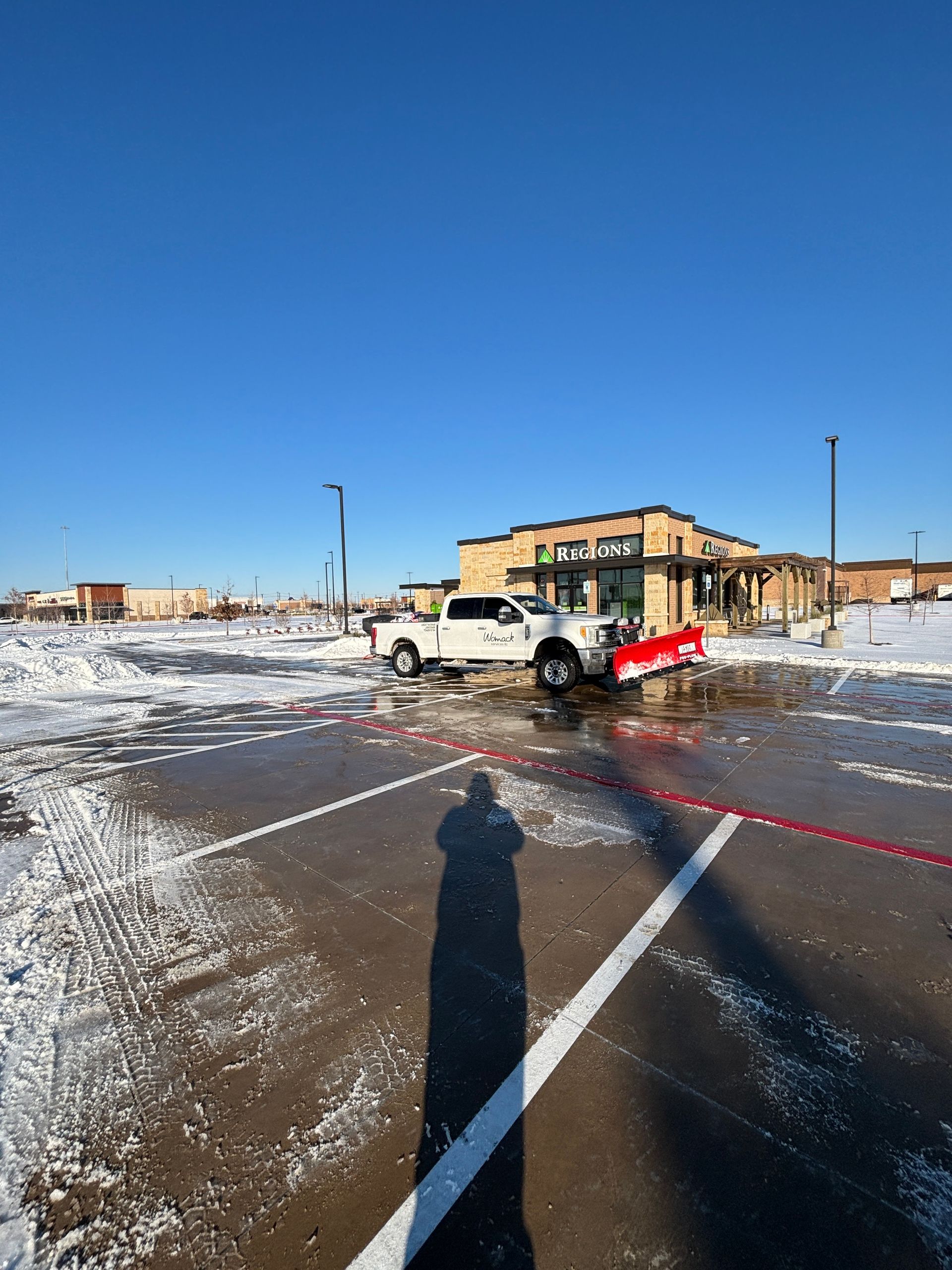 Snowy parking lot with a white truck towing a red snowplow near a building under a clear blue sky