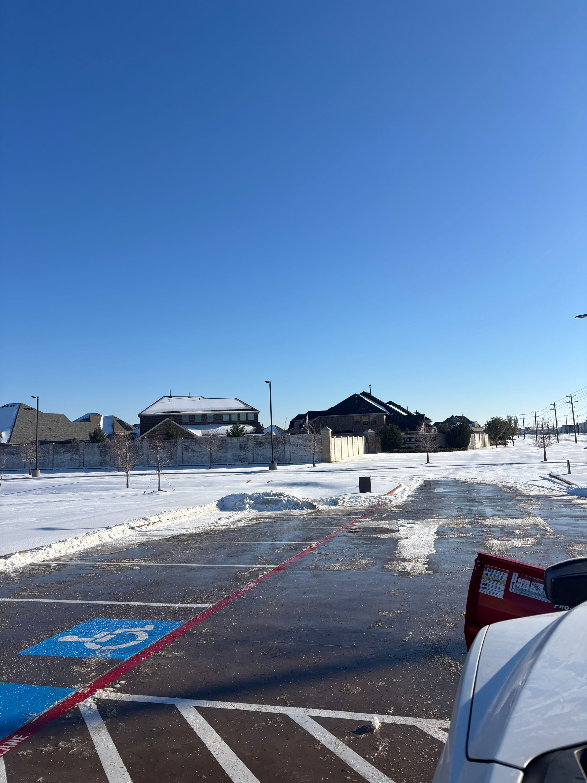 Snowy parking lot with icy pavement, red lines, and houses under a clear blue sky