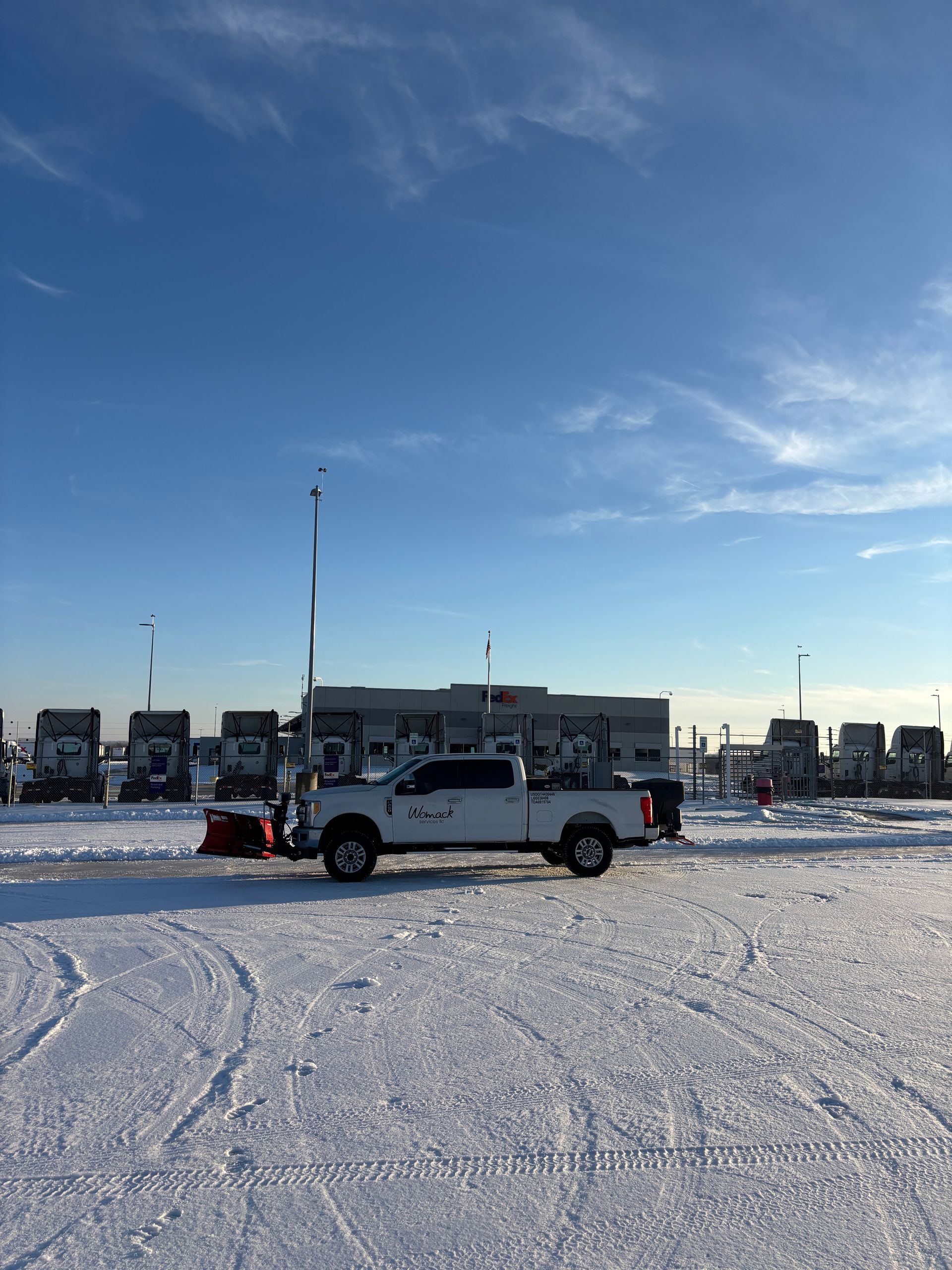 Pickup truck plowing snow in a bright, snowy parking lot with a row of vehicles and buildings behind it