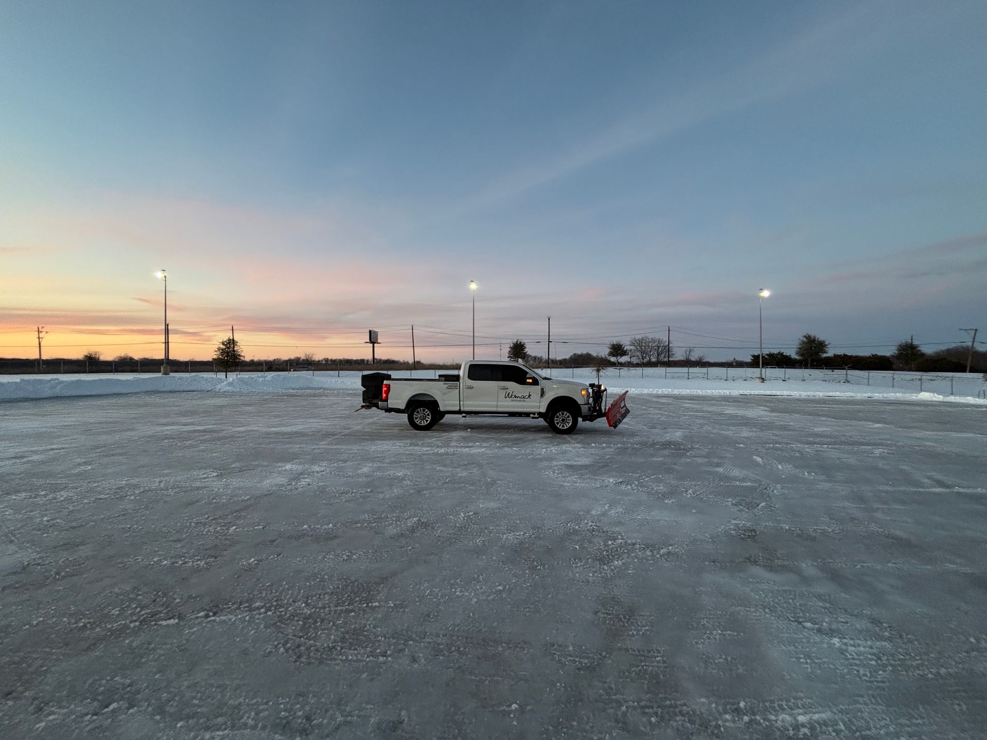 Pickup truck on a snowy road at sunset with a person standing nearby and streetlights in the distance