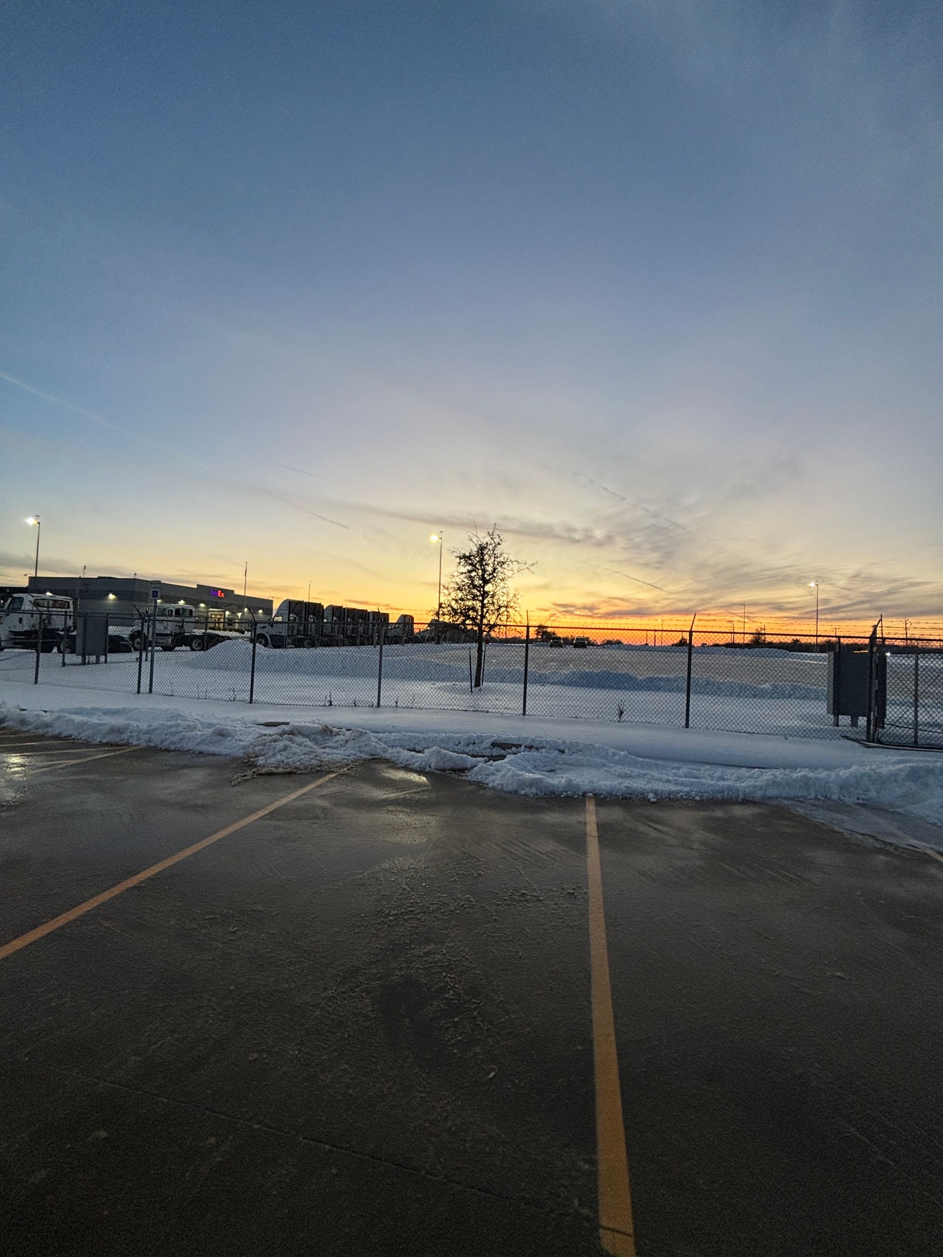 Snowy parking lot at sunset with a fence and distant buildings under a blue-orange sky
