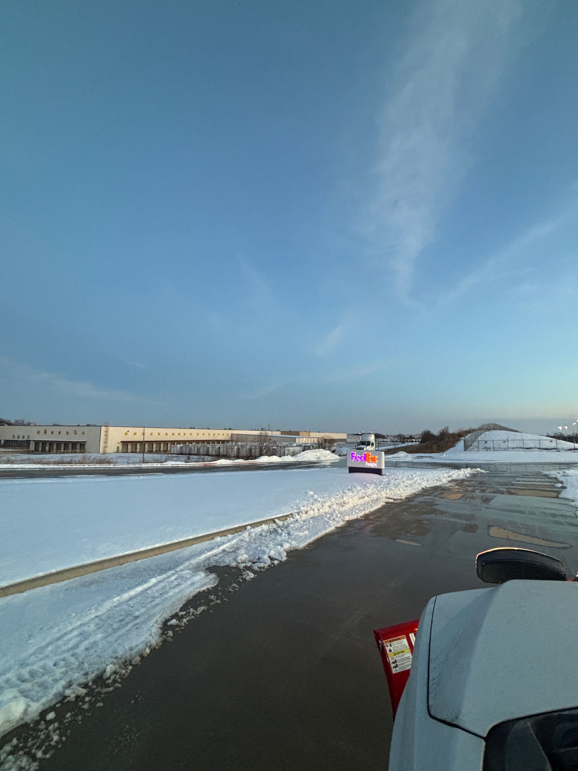 Snowy runway beside water, with a red-and-blue vehicle driving ahead under a blue sky.