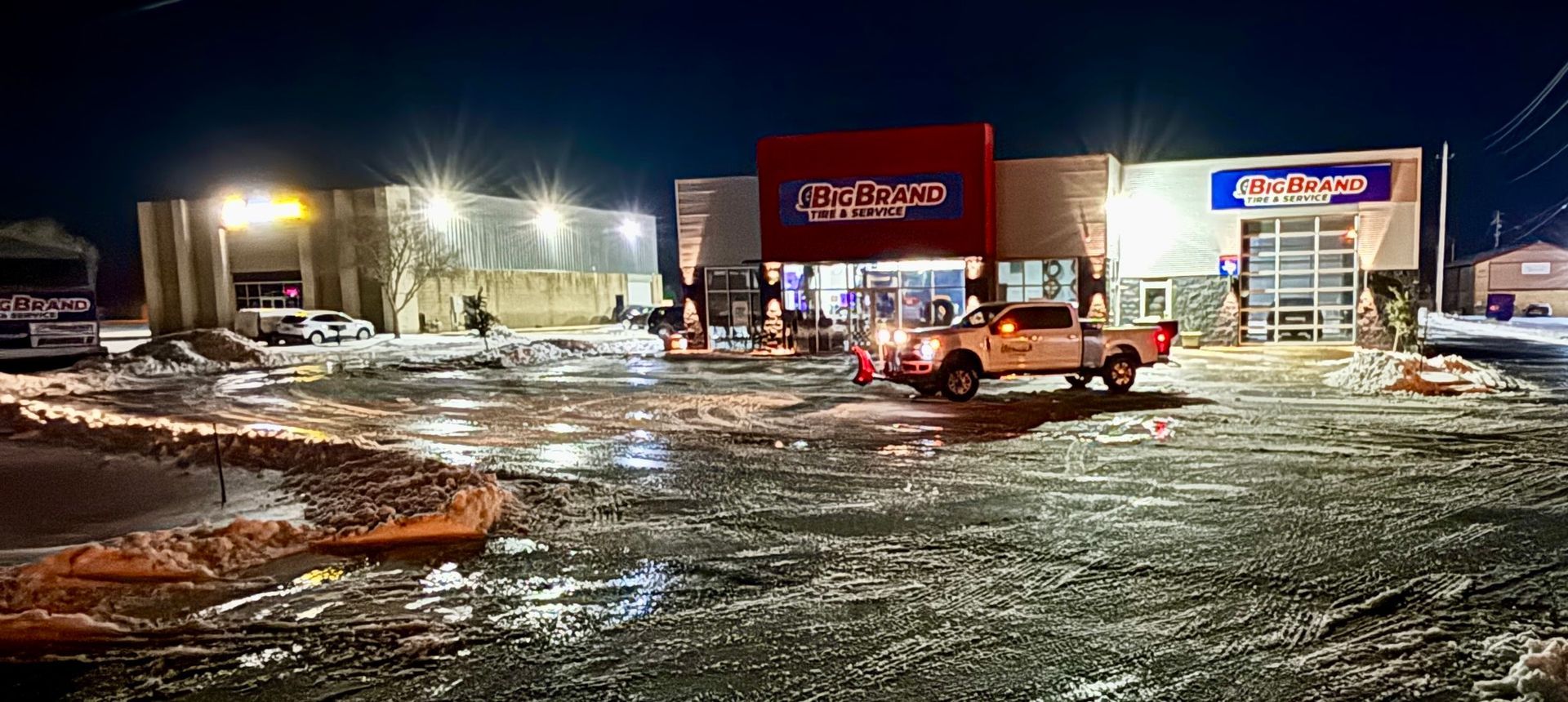 Snowy parking lot outside a red storefront at night, with cars and bright lights reflected on wet ground