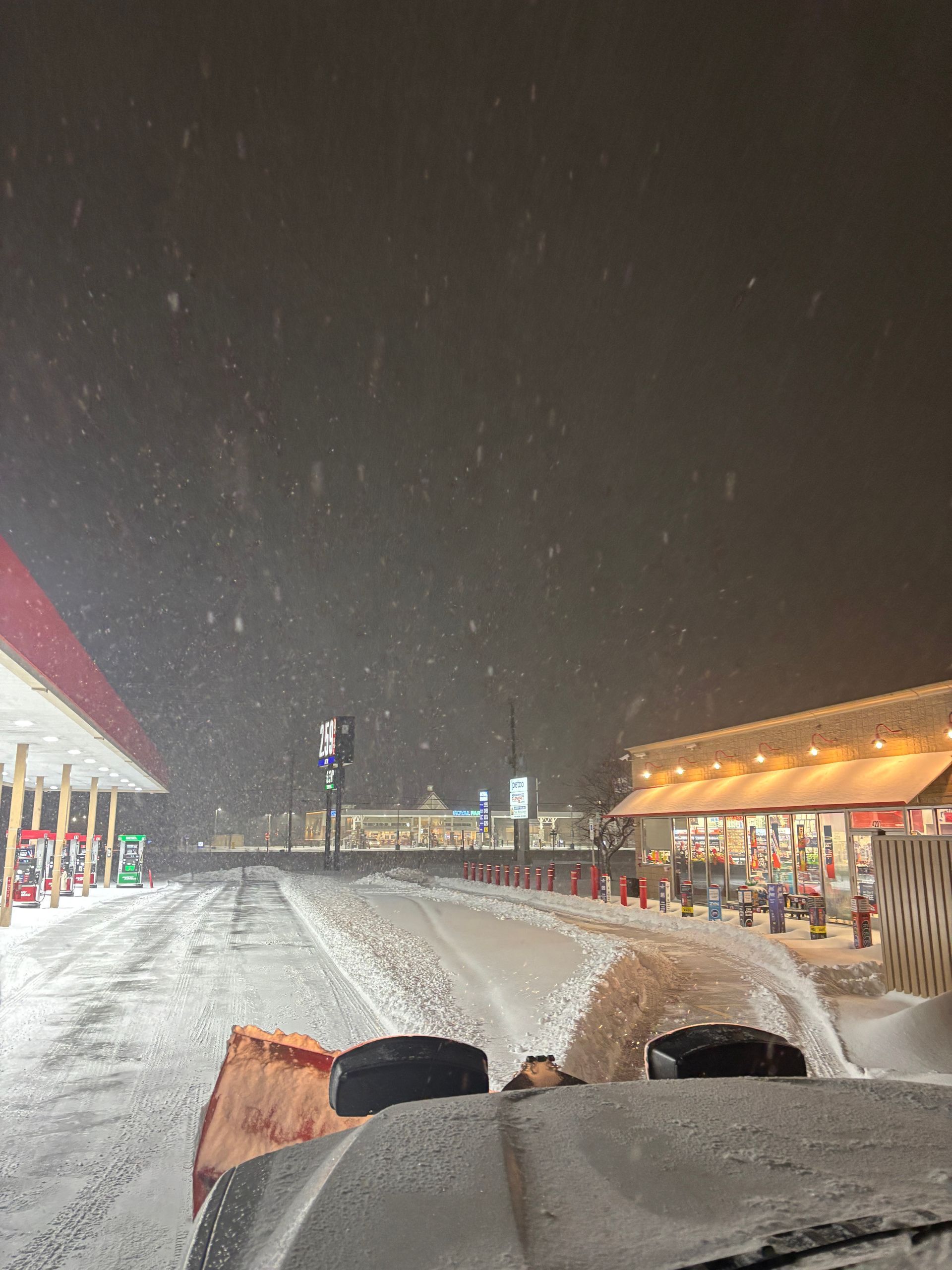 Snowy night at a lit outdoor market, viewed from a moving vehicle on a slushy road.