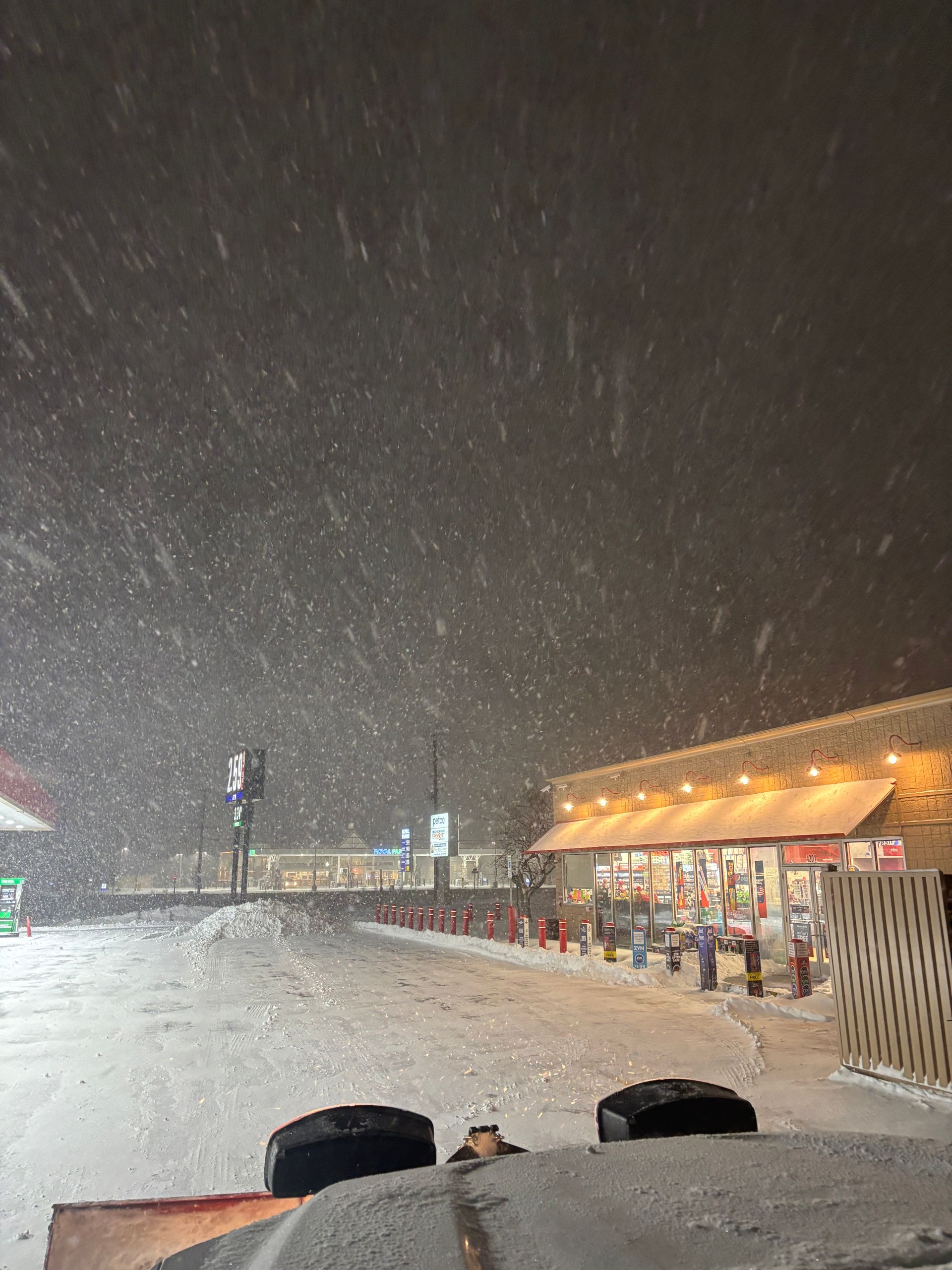 Snowy night scene outside a lit storefront with people standing under the awning.