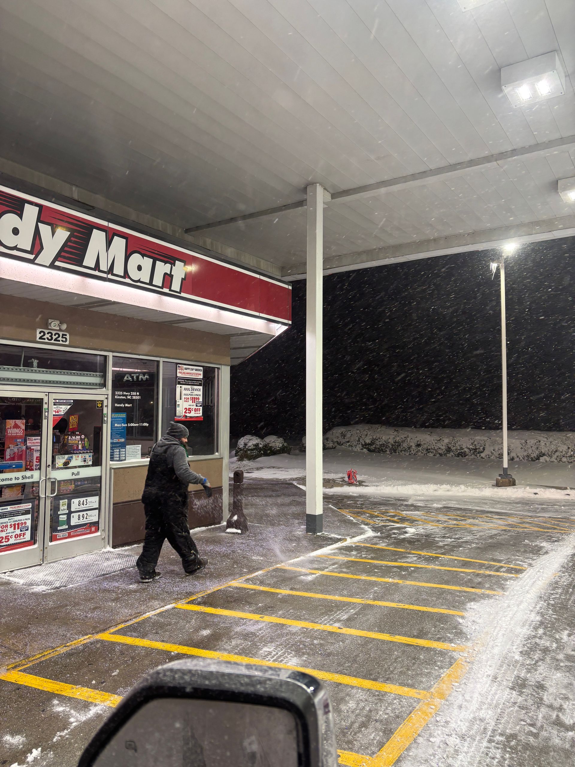 Person walking under a gas station canopy in snowy night conditions with lit storefront and parking lot lines
