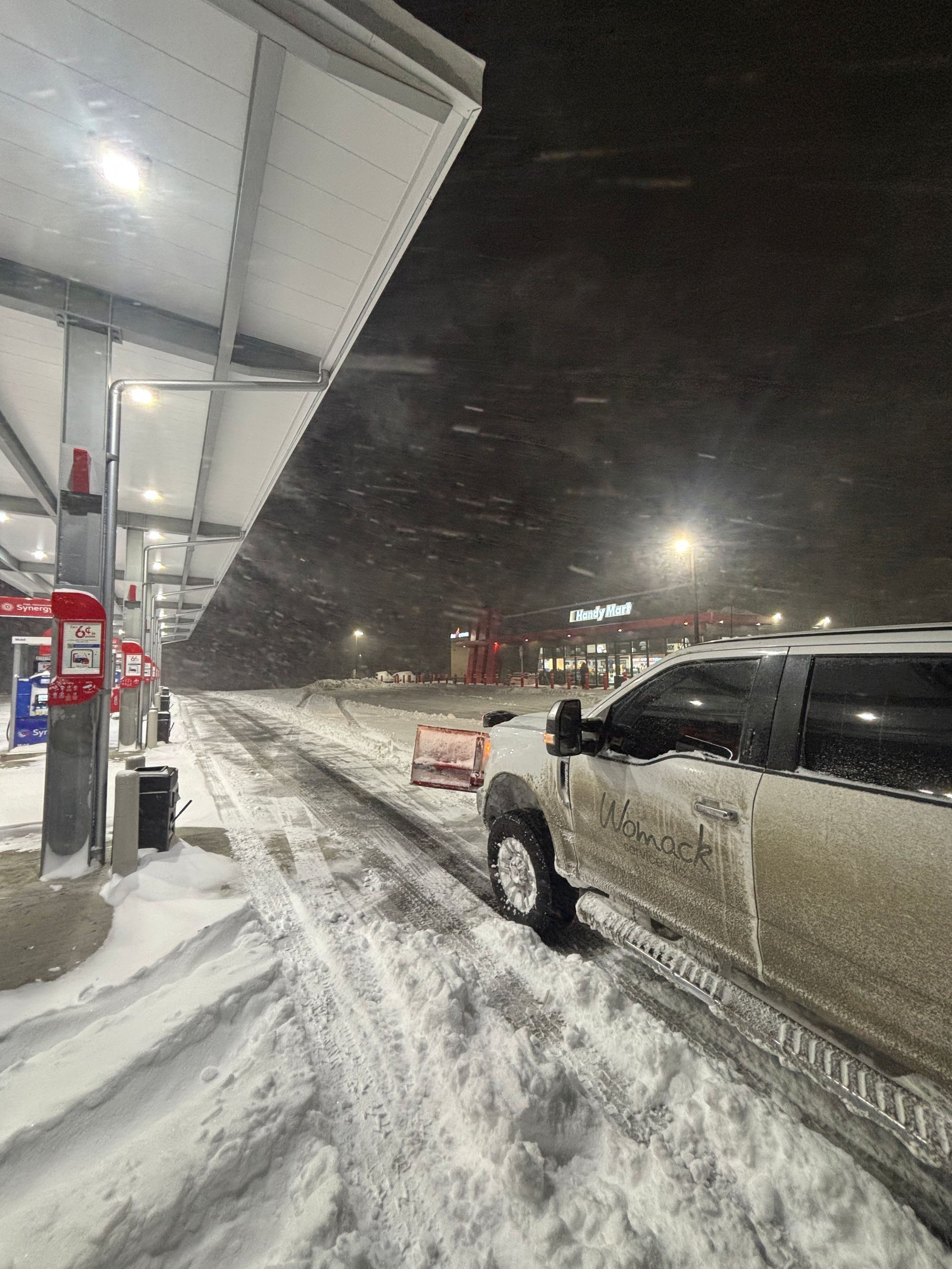 Snowy gas station at night with a parked pickup truck and heavy snowfall.