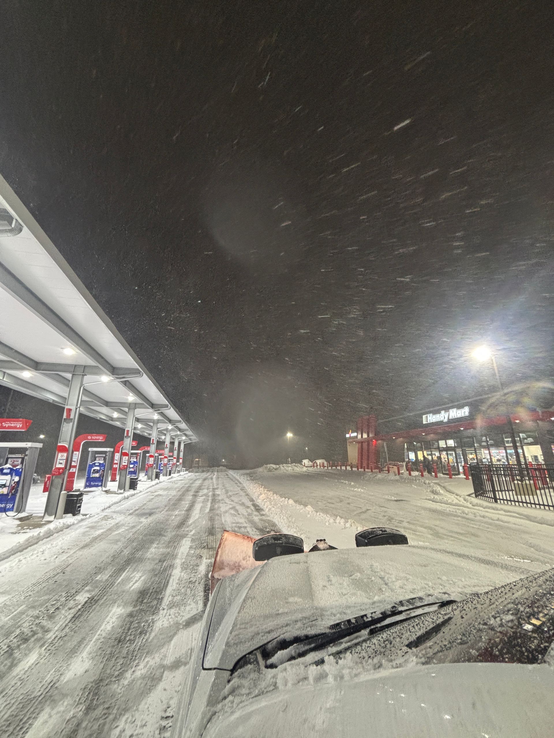 Snowy gas station at night with cars covered in snow and heavy snowfall visible in the air