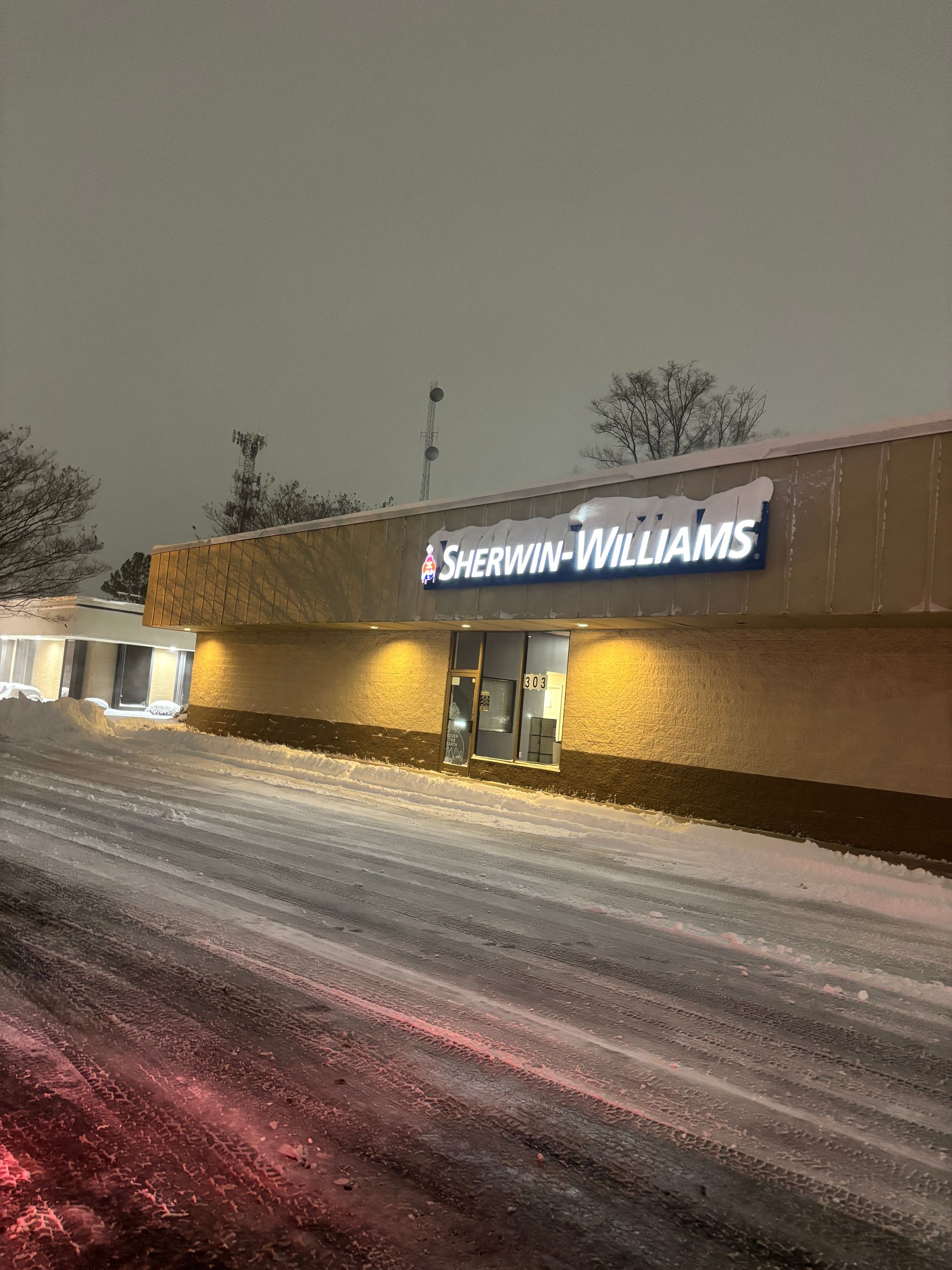 Snow-covered Sherwin-Williams store at dusk, with illuminated sign and snowy parking lot.