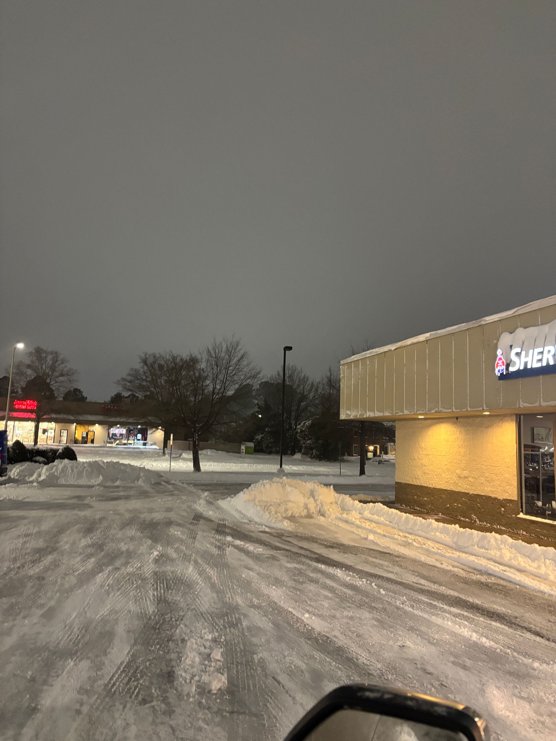 Snowy parking lot at dusk beside a lit storefront and piled snowbanks.